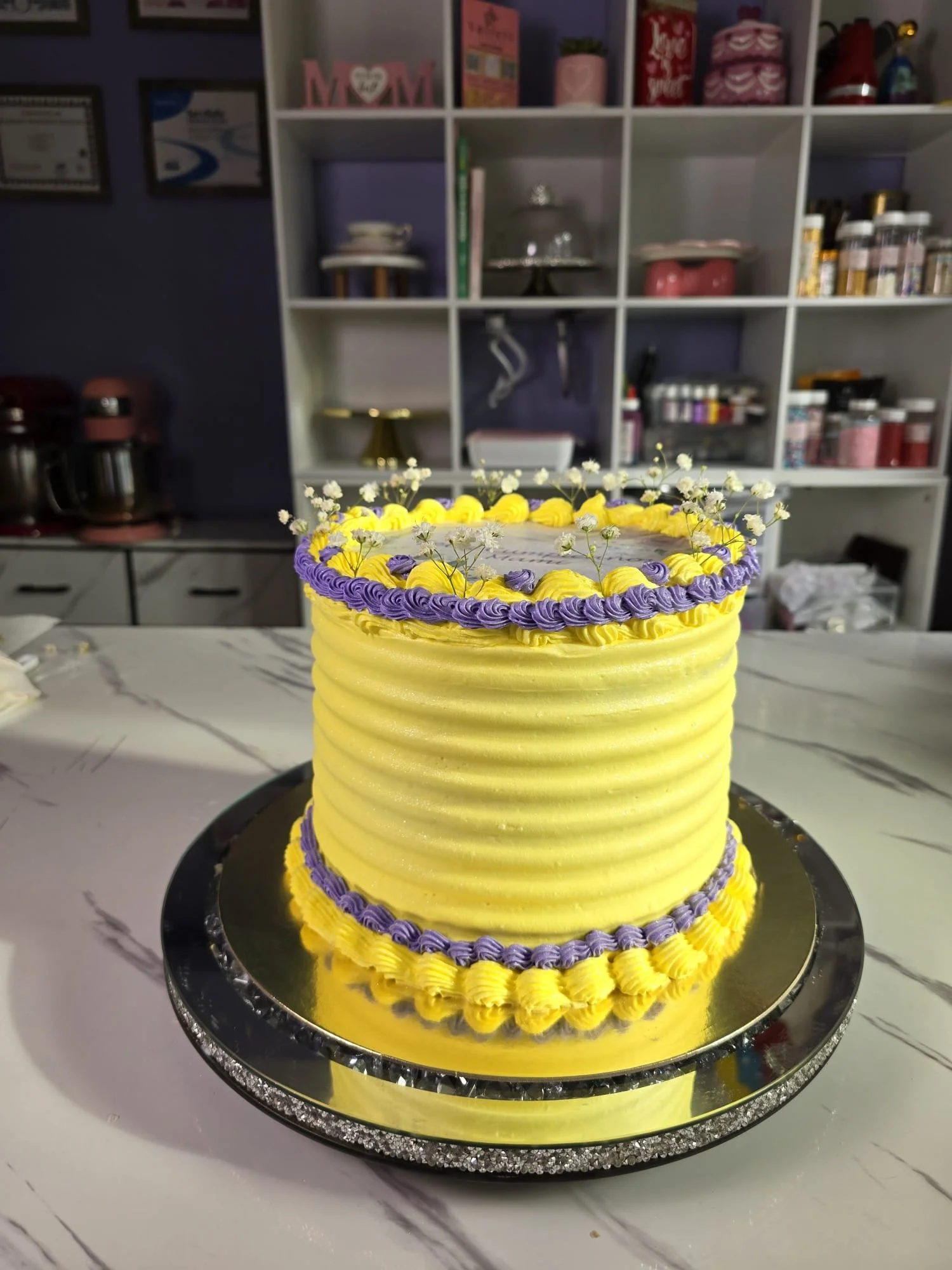 Pastel de cumpleaños de dos pisos en colores amarillo y morado, decorado con flores blancas y en un plato reflectante en una mesa de mármol, con una estantería blanca en el fondo.