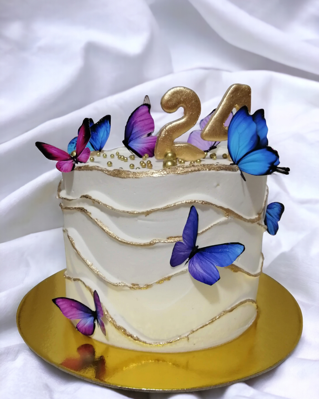 Pastel de cumpleaños blanco decorado con mariposas moradas y azules, números dorados 24, y pequeñas perlas doradas. Está en un plato dorado sobre tela blanca.