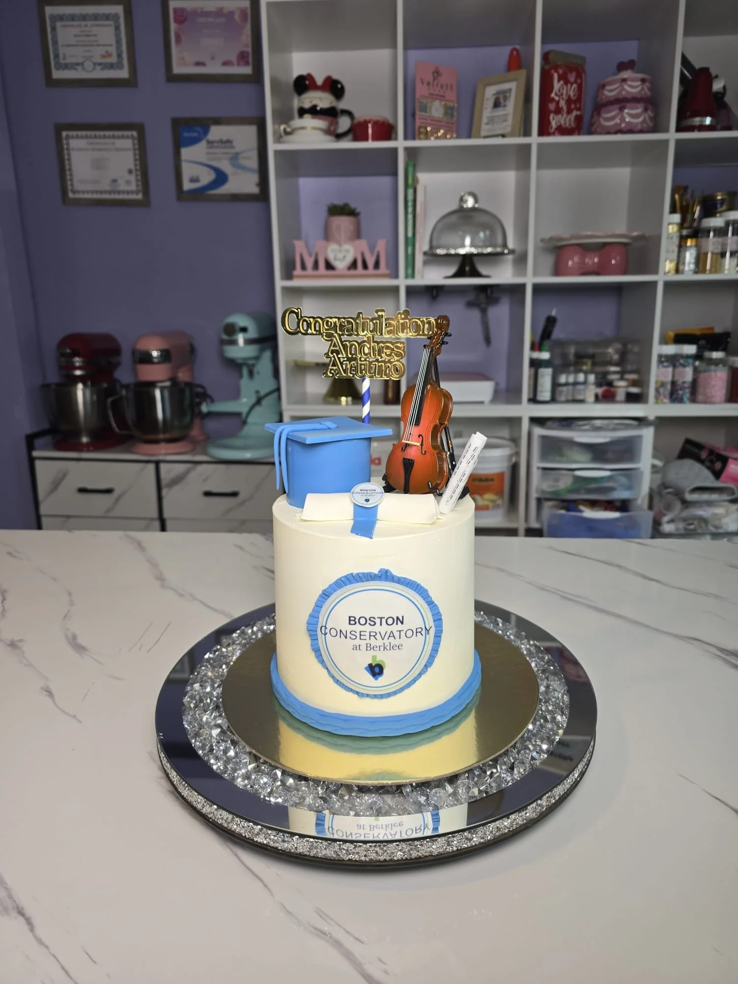Pastel de cumpleaños con motivo institucional, decoraciones en azul y blanco, con adornos musicales y una felicitación de graduación, en una mesa de mármol con fondo de estantería con certificados, libros y objetos decorativos.