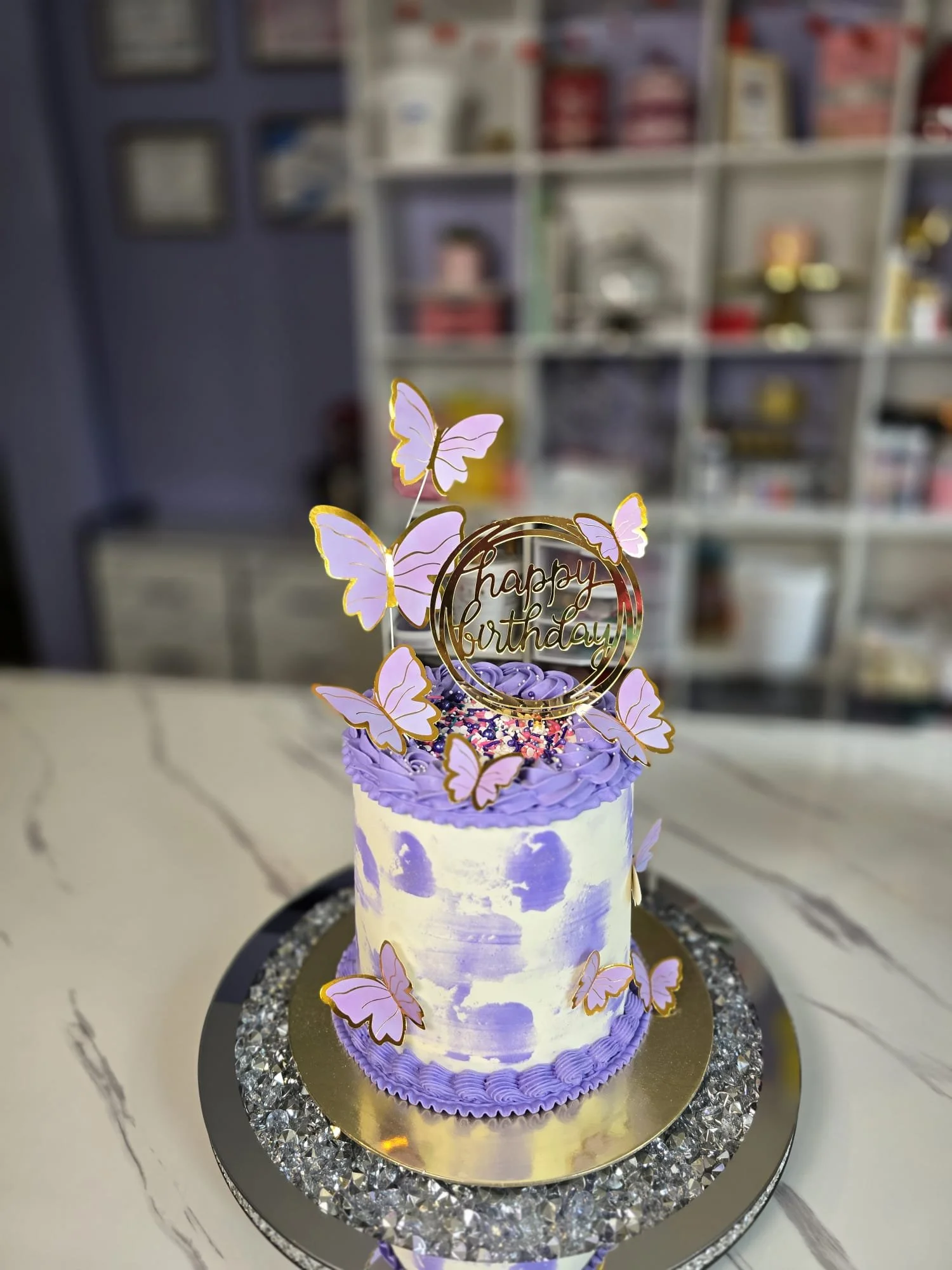 Pastel de cumpleaños con decoraciones de mariposas moradas y un cartel que dice 'Happy Birthday' sobre un plato brillante.