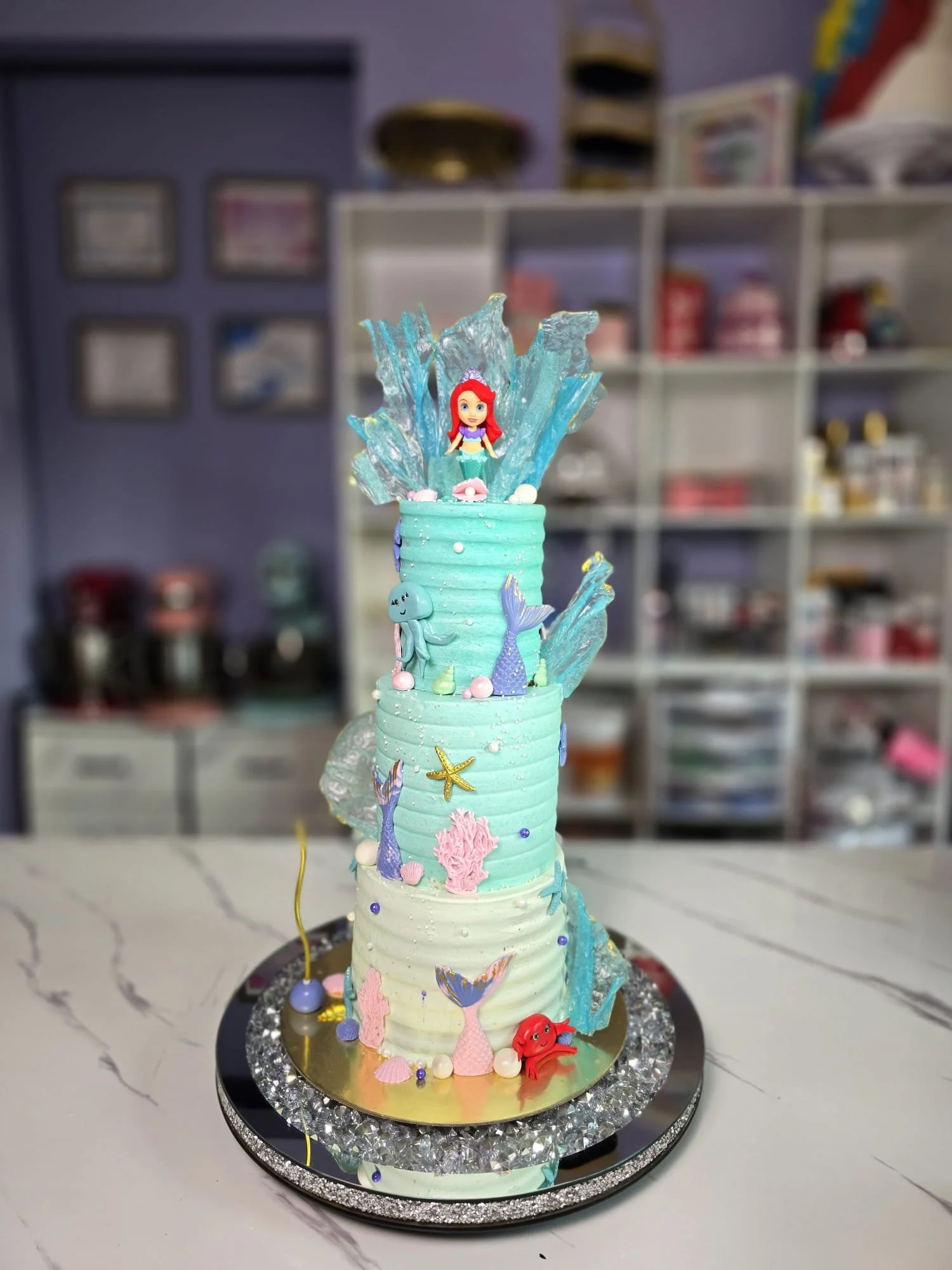 Pastel de cumpleaños con decoración marina, incluyendo figuras de la sirenita Ariel, estrellas de mar, conchas, fragmentos de hielo y decoraciones en tonos azules y rosados.