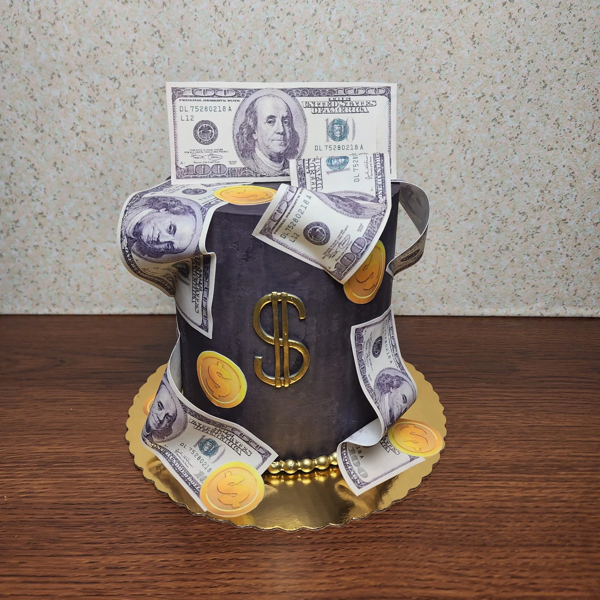 Pastel de cumpleaños decorado con billetes de cien dólares y monedas de oro, con un símbolo de dólar en el centro, sobre una base dorada en una mesa de madera.