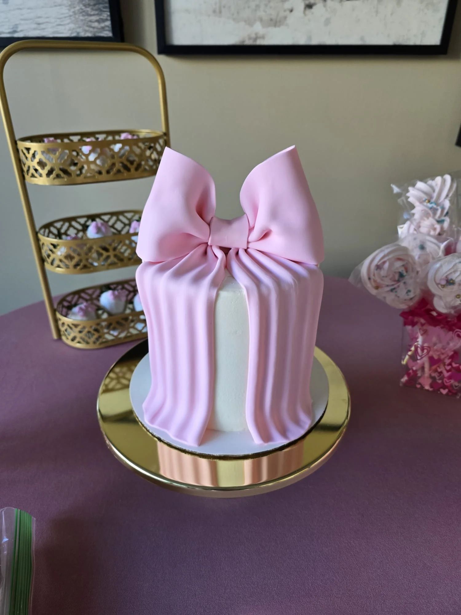 Pastel de cumpleaños decorado con una gran cinta rosa que parece un lazo, colocado sobre un plato dorado. Al fondo, hay una bandeja dorada con dulces decorativos y una taza con dulces en forma de nube con colores pastel.