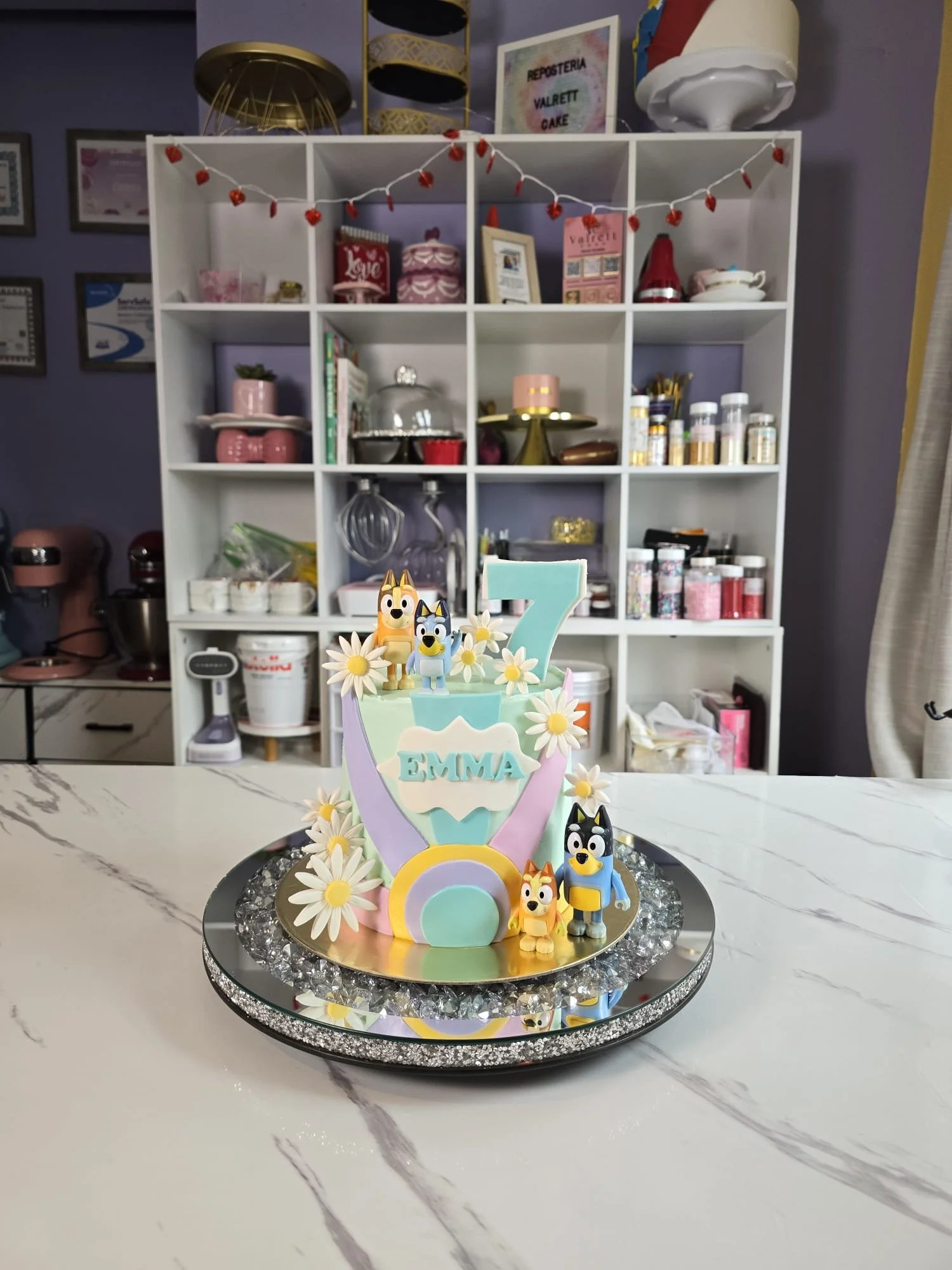 Pastel de cumpleaños decorado con temática de perros y flores, con el número 7 y la palabra 'Emma' en el frente, colocado sobre un plato plateado con piedras decorativas.