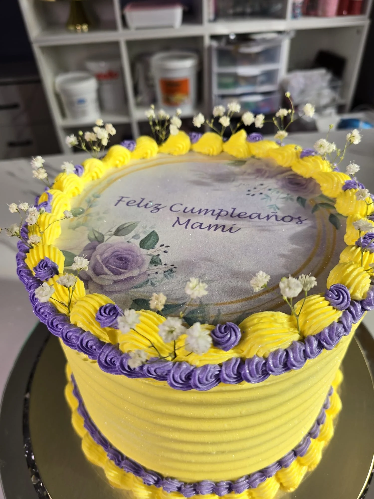 Pastel de cumpleaños para mamá con decoraciones en colores amarillo y morado, con flores blancas y mensaje que dice 'Feliz Cumpleaños Mamá' en la parte superior.