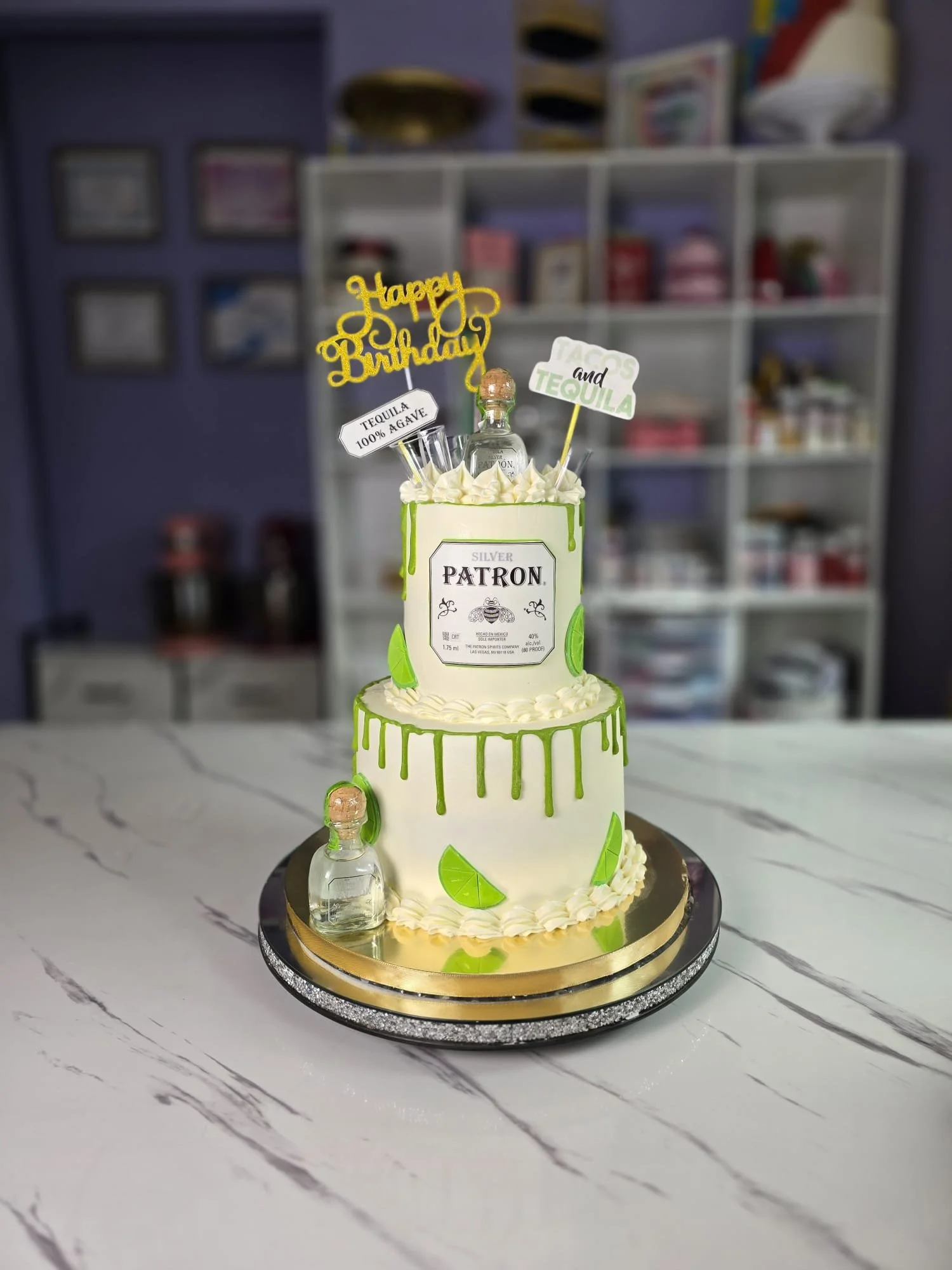 Pastel de cumpleaños decorado con temática de tequila, con pequeñas botellas de tequila y limones de fondant, en un entorno de cocina o sala con estantes y objetos en el fondo.