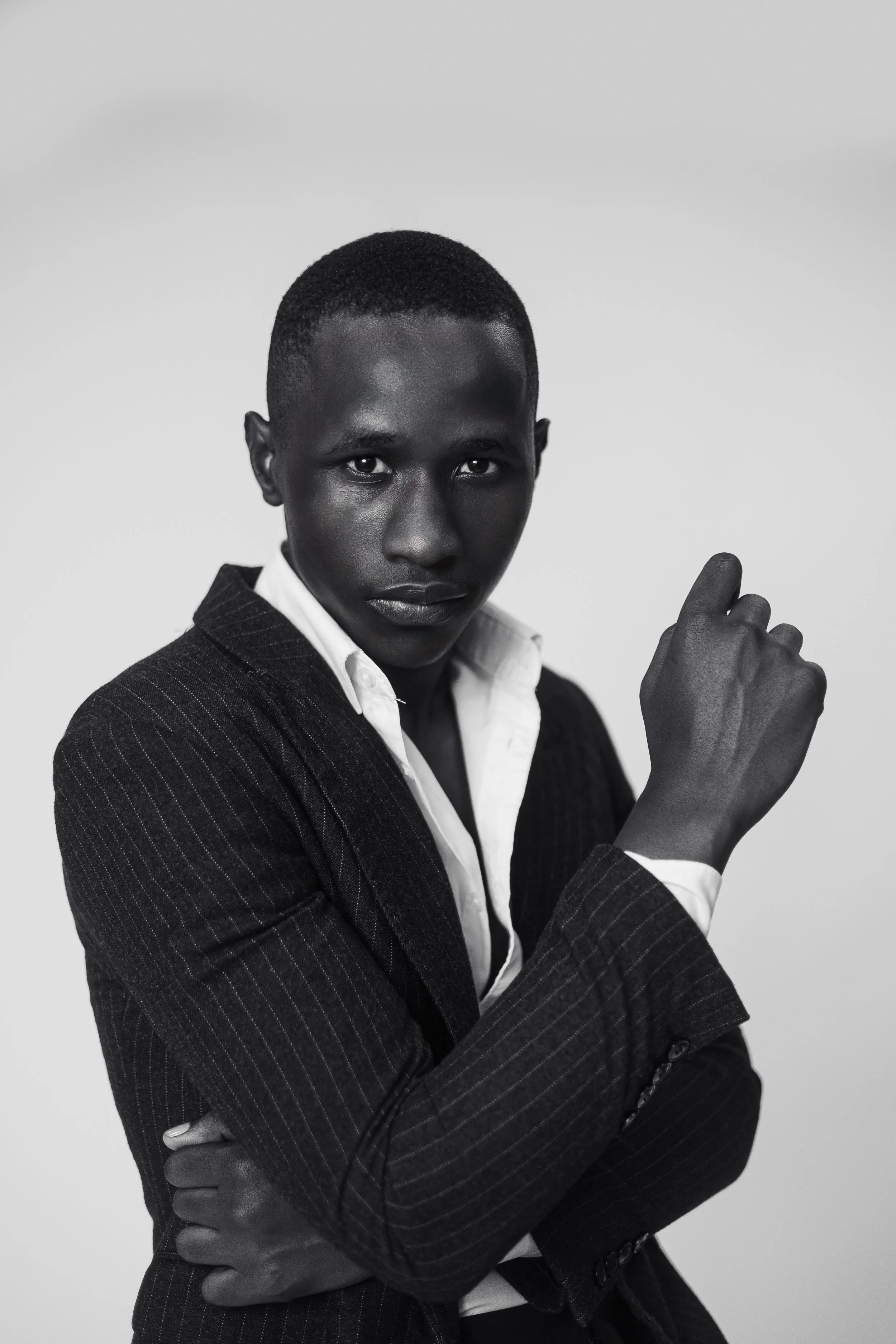 Black and white photo of a young man wearing a pinstripe suit and white shirt, with short hair, looking directly at the camera, crossing his arms, against a plain background.