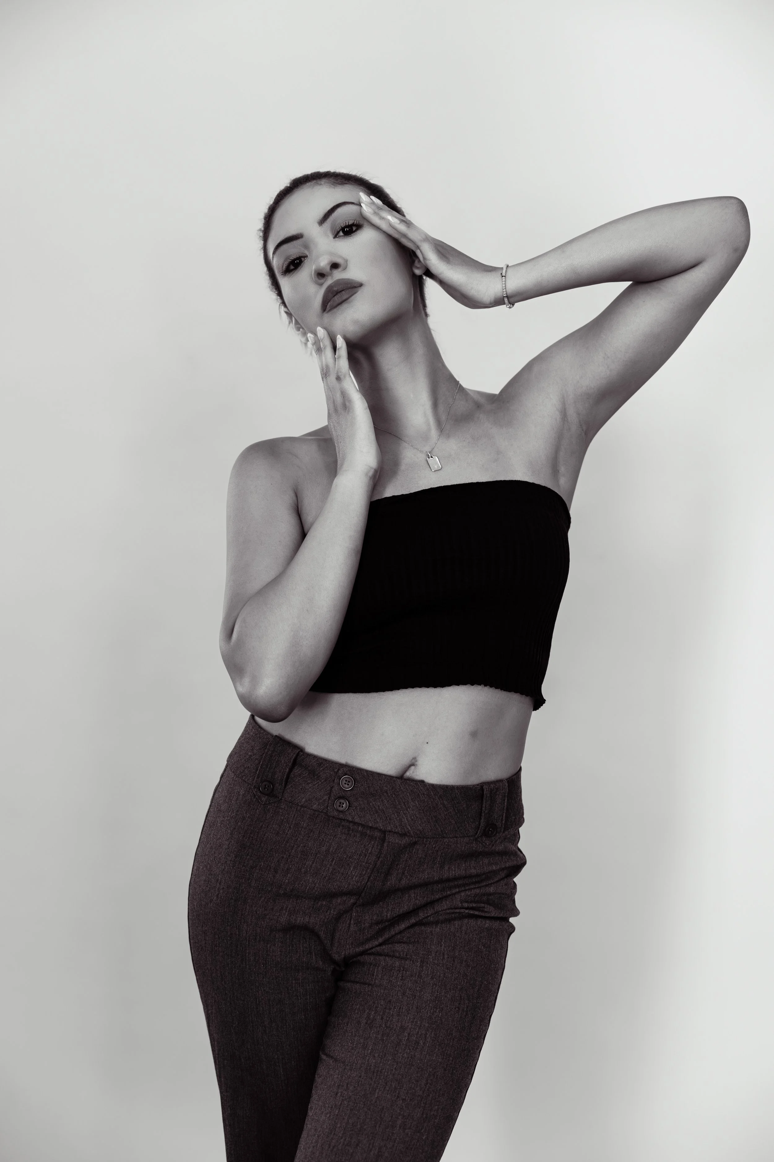 Black and white photograph of a woman with short hair, wearing a strapless top and high-waisted pants, posing with her hands on her face and neck, against a plain background.