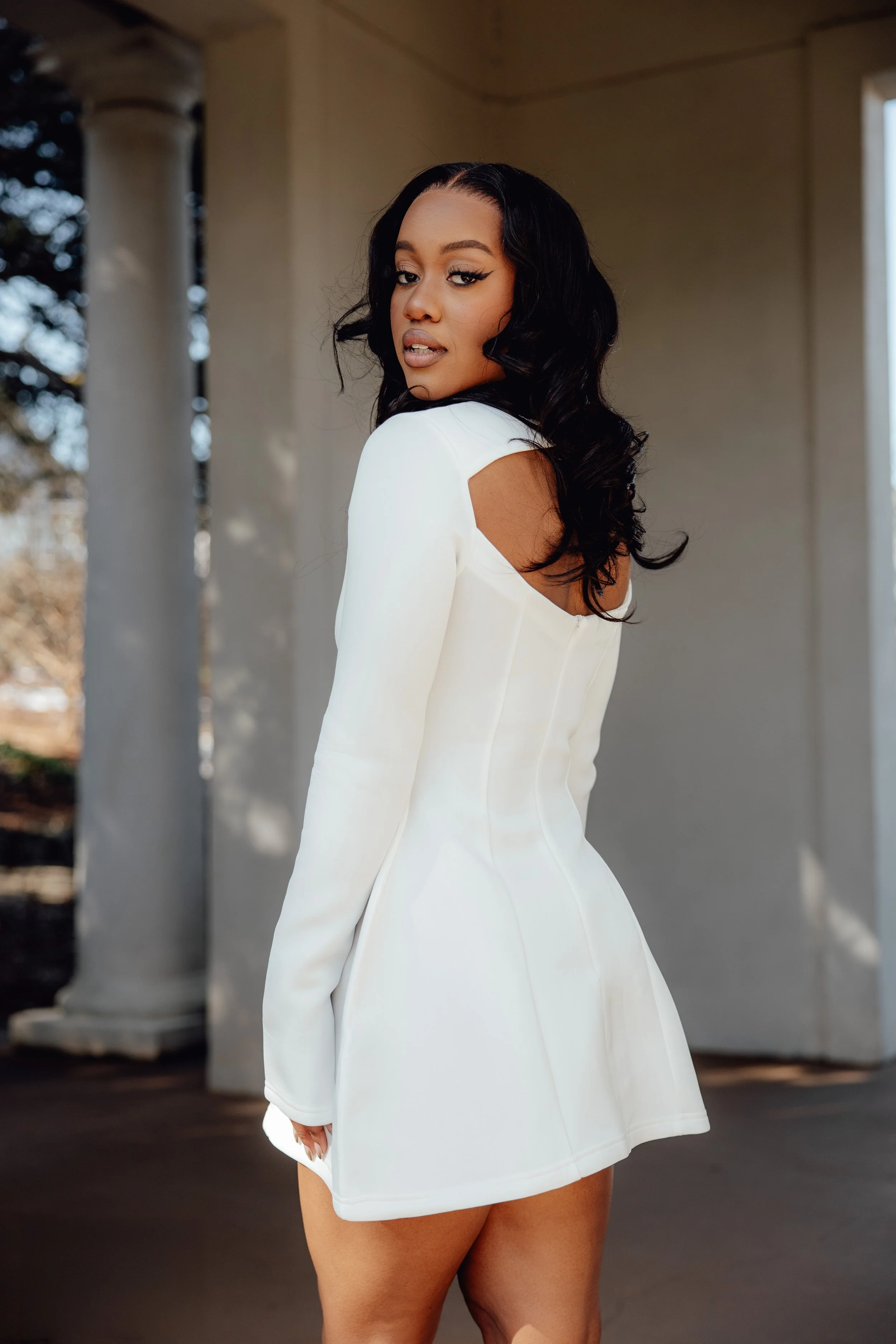 A woman with long black hair wearing a white dress with cut-out shoulders, standing outdoors near a white column, with trees and a bright sky in the background.