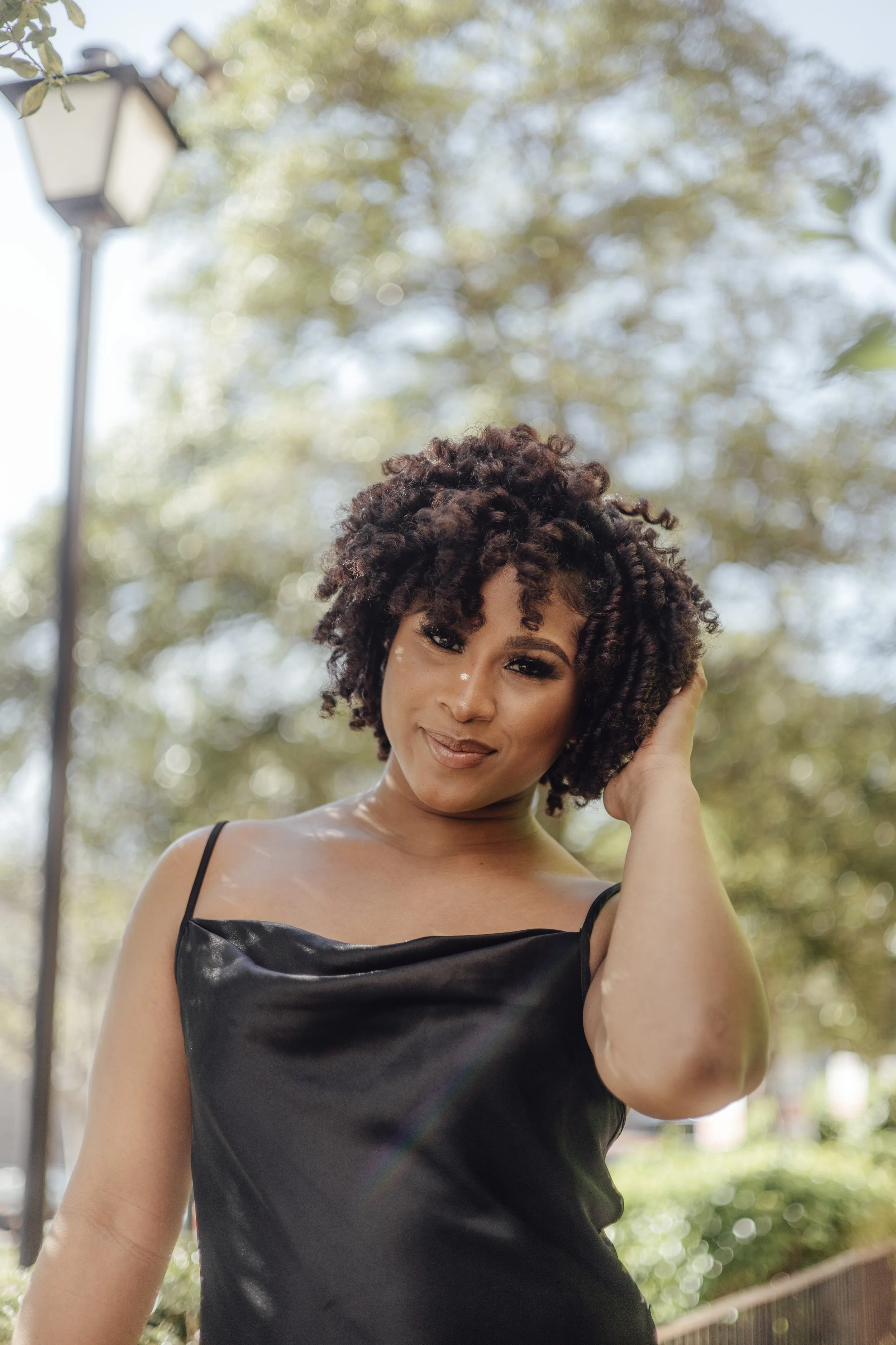 A woman with curly dark hair wearing a black satin slip dress outdoors, touching her hair, with trees and a lamp post in the background.
