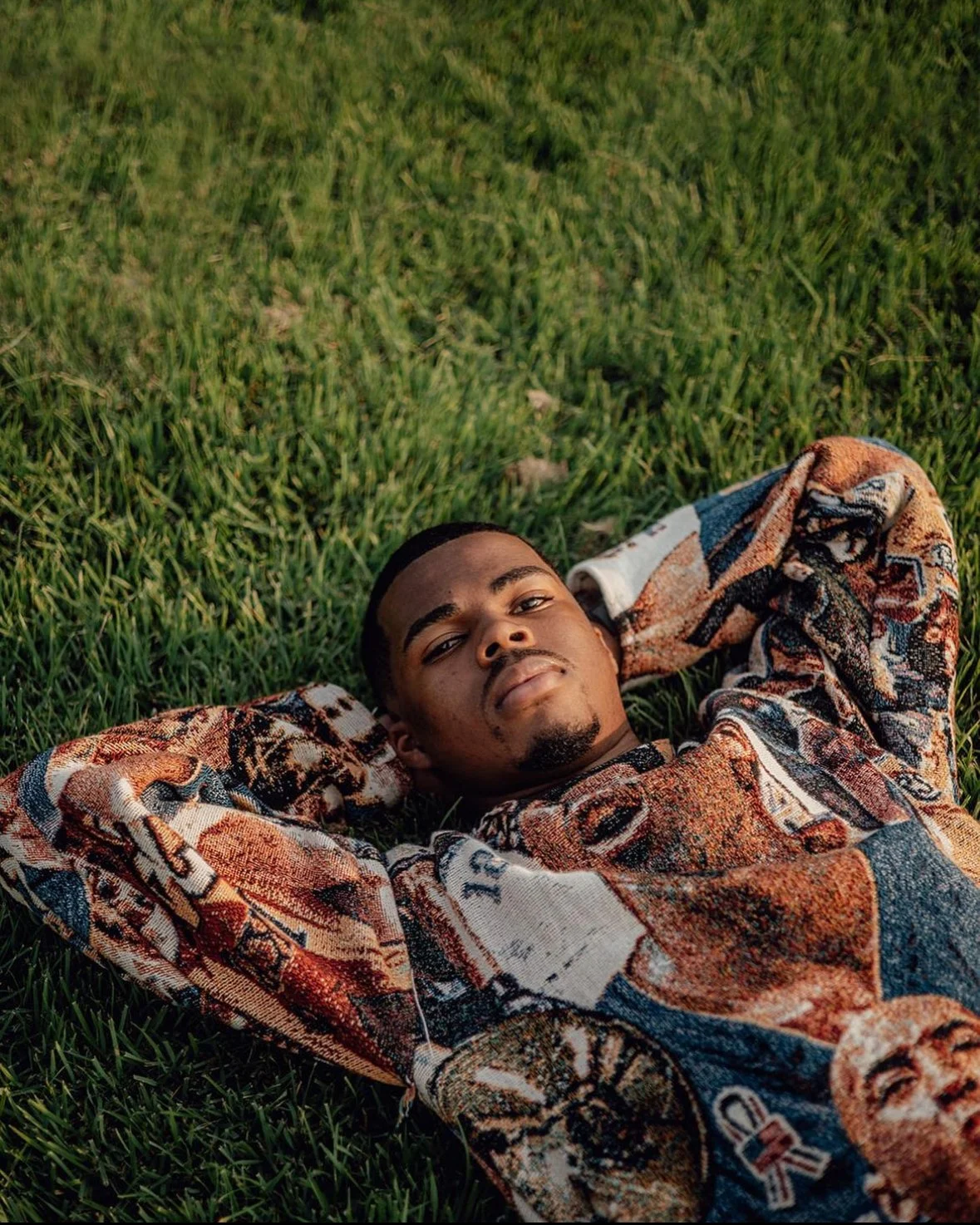 A young man lies on his back on green grass, with his arms resting behind his head, looking at the camera.