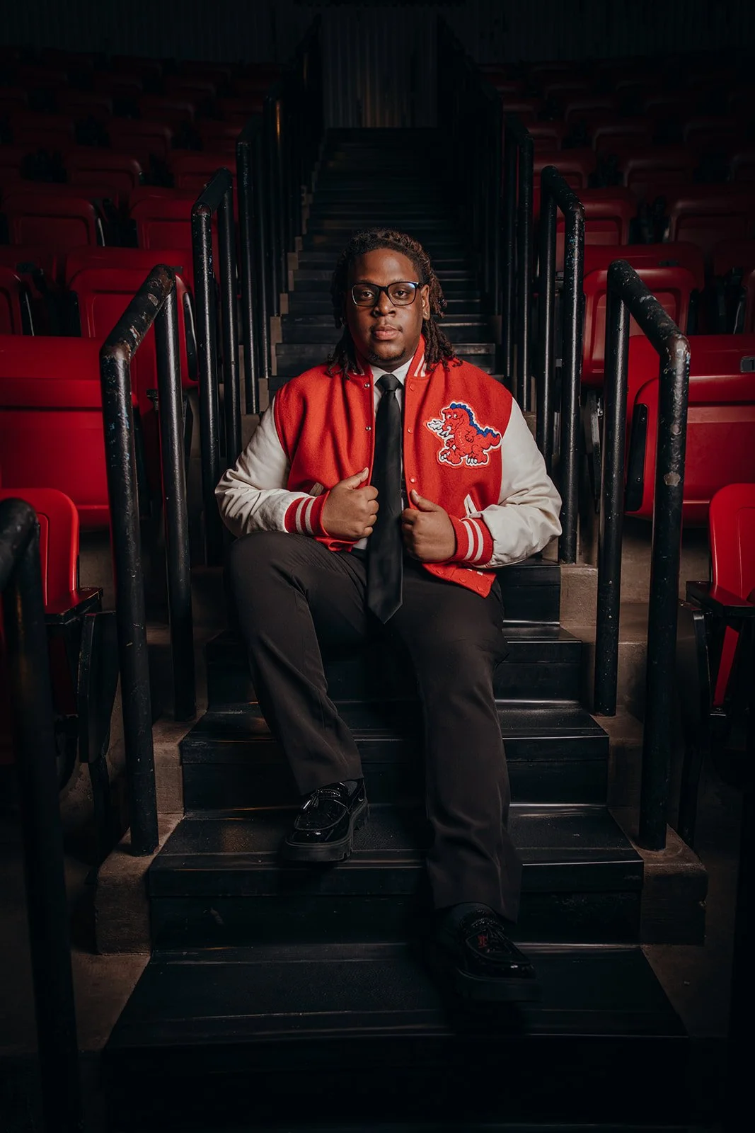 A man with glasses and dreadlocks sitting on black stadium stairs, wearing a red and white varsity jacket with a lion patch, black pants, black shoes, white shirt, and black tie, in an empty arena with red seats.