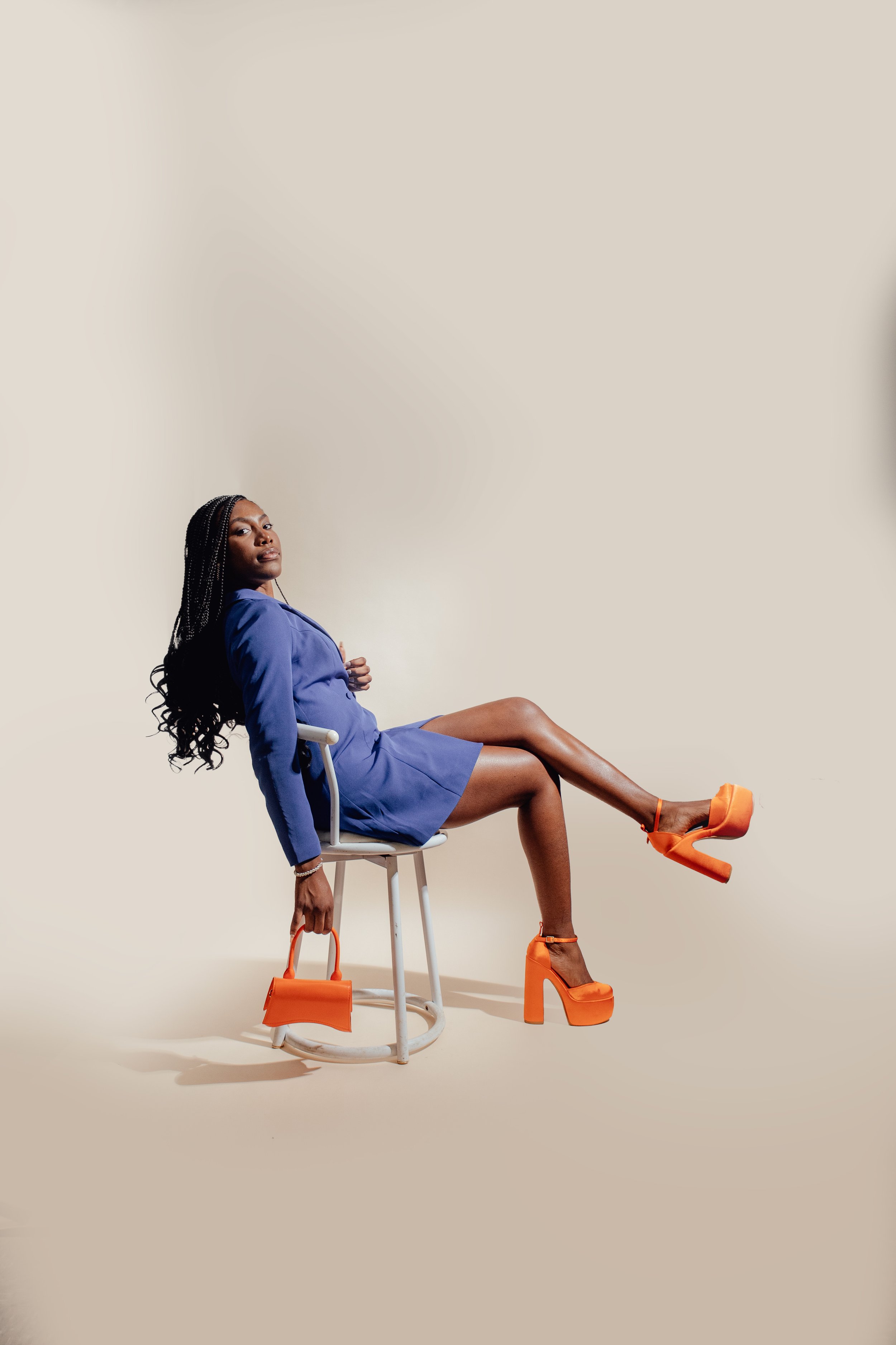 A woman with braided hair, wearing a blue blazer and orange platform heels, sitting on a chair with her legs crossed and holding an orange handbag.