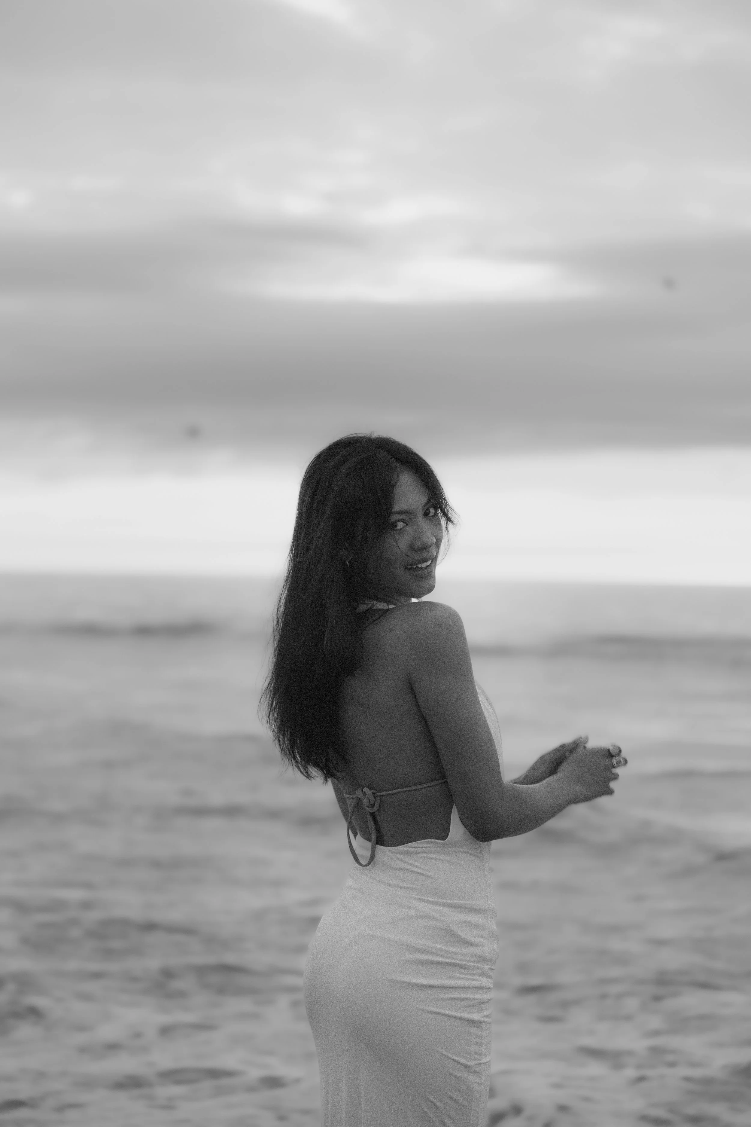 A woman standing on the beach with the ocean and cloudy sky in the background, wearing a light-colored dress and looking back at the camera.