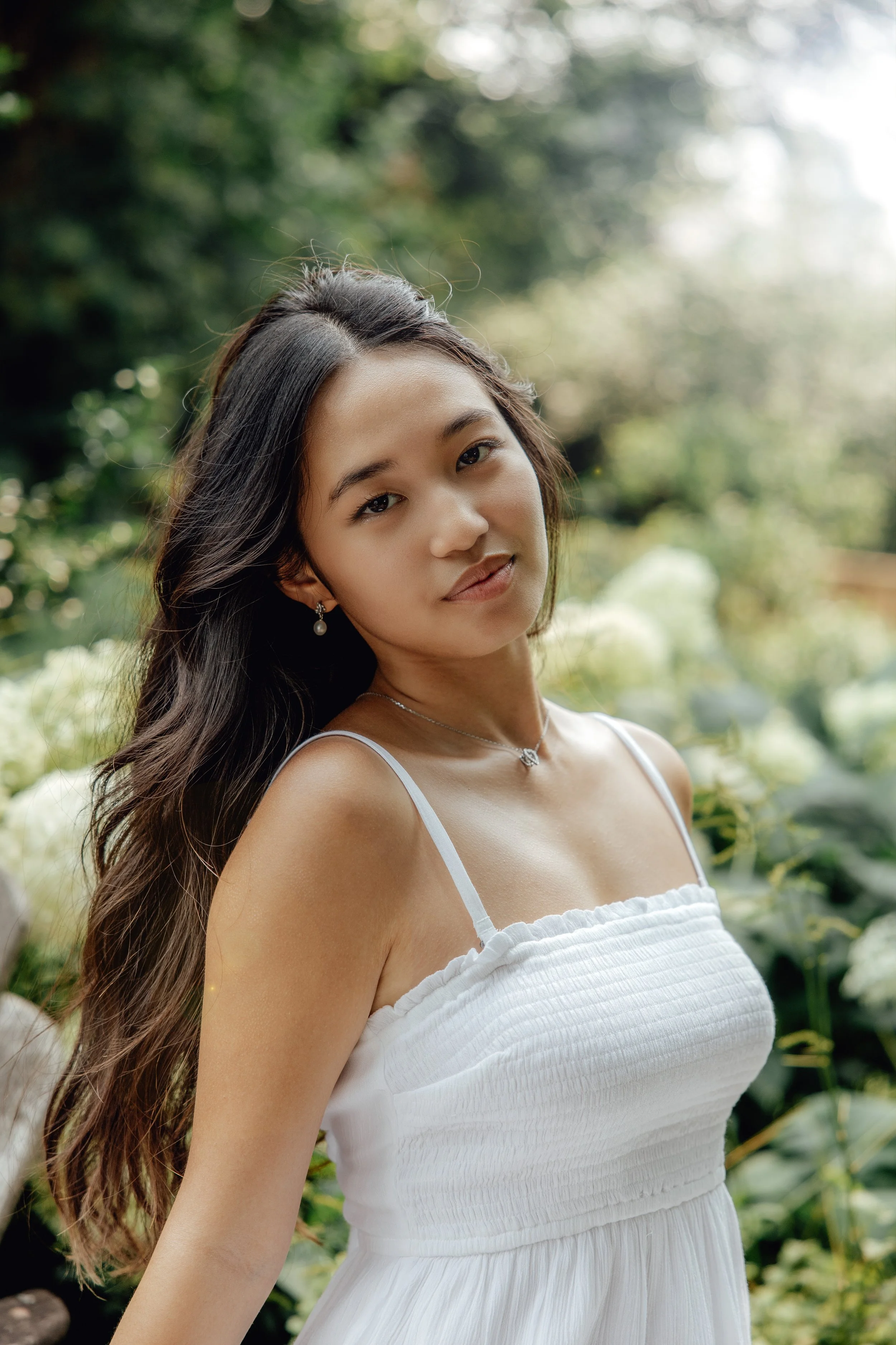 A young woman with long dark hair in loose waves, wearing a white sleeveless dress and pearl jewelry, standing outdoors among greenery with sunlight filtering through trees.