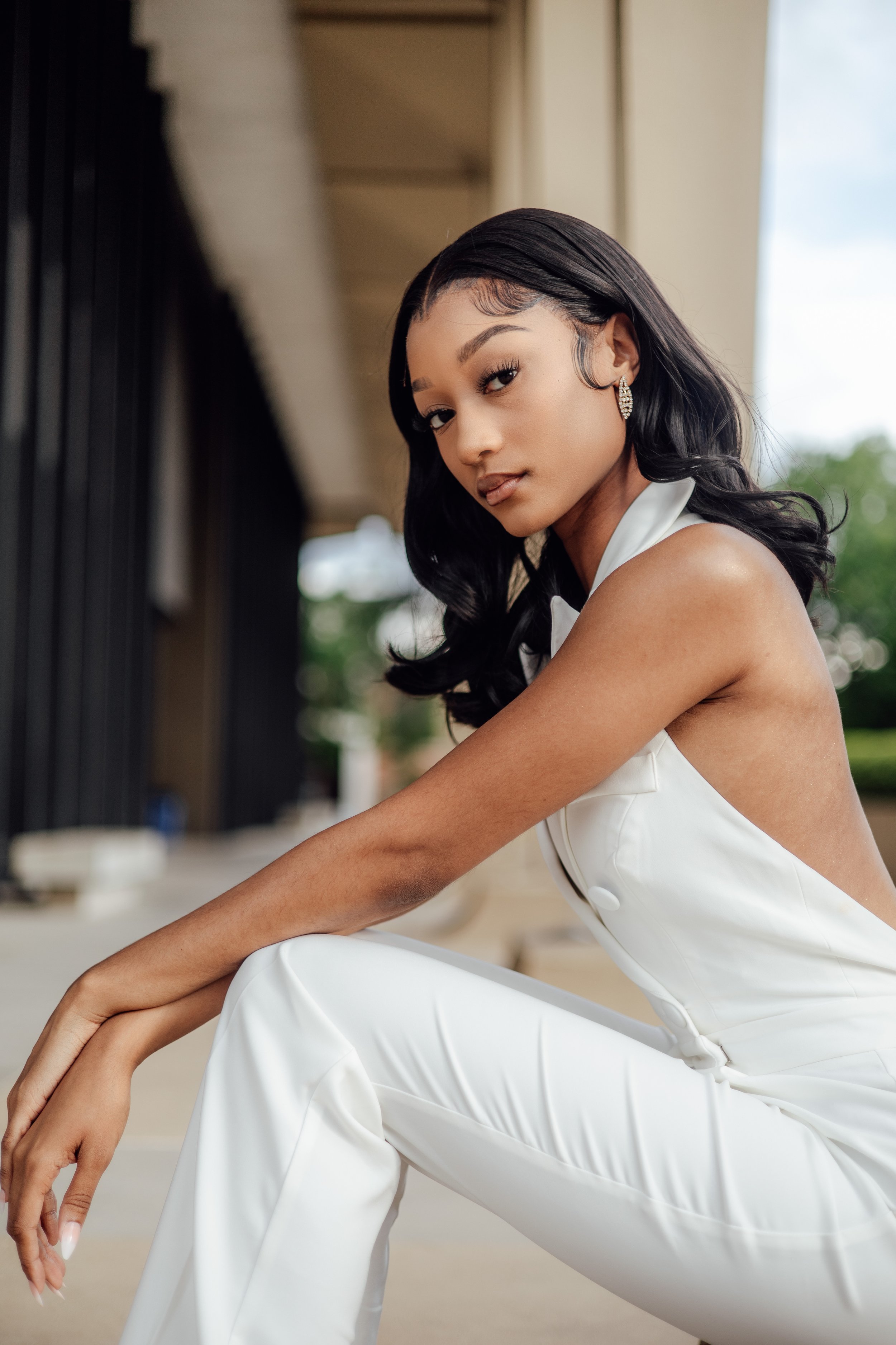 A young woman with black wavy hair and light makeup, wearing a white sleeveless outfit with a high collar, sitting outdoors near a building with a blurred background of greenery and sky.
