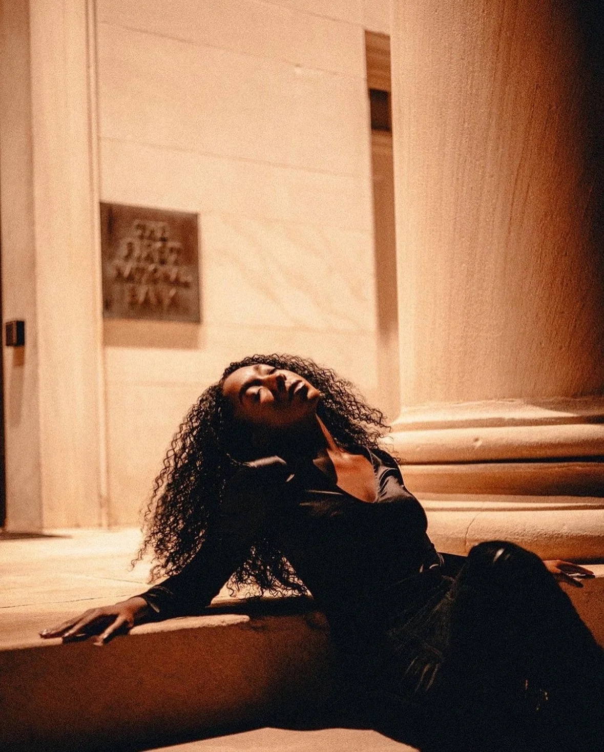 A woman with curly hair and dark clothing sitting on the floor, leaning against a ledge, with her eyes closed and head tilted back in a relaxed or contemplative pose, in a dimly lit indoor space with wooden walls.