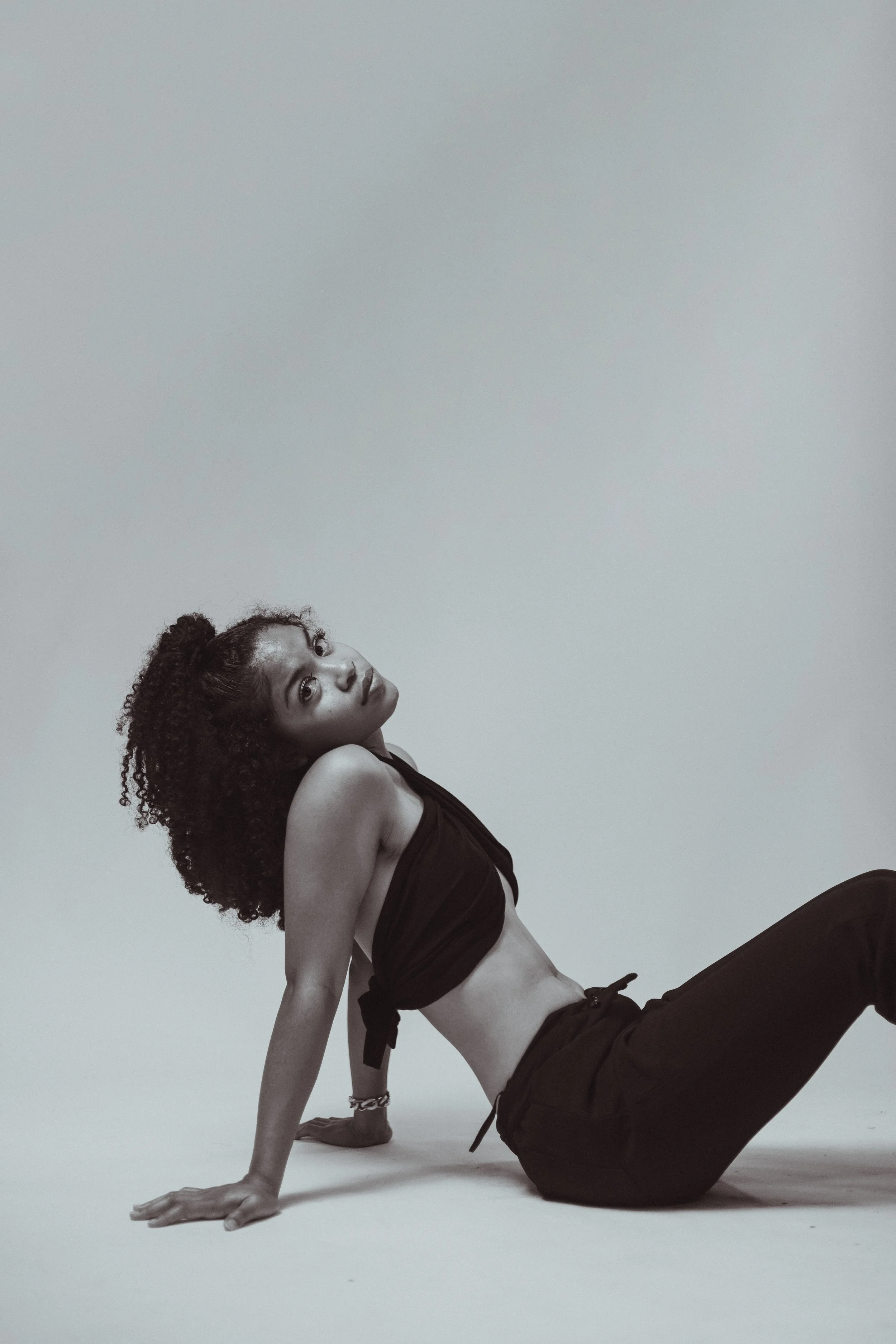 A woman with curly hair poses in a black tank top and shorts, sitting on the ground with her back arched and head tilted back, against a plain light gray background.