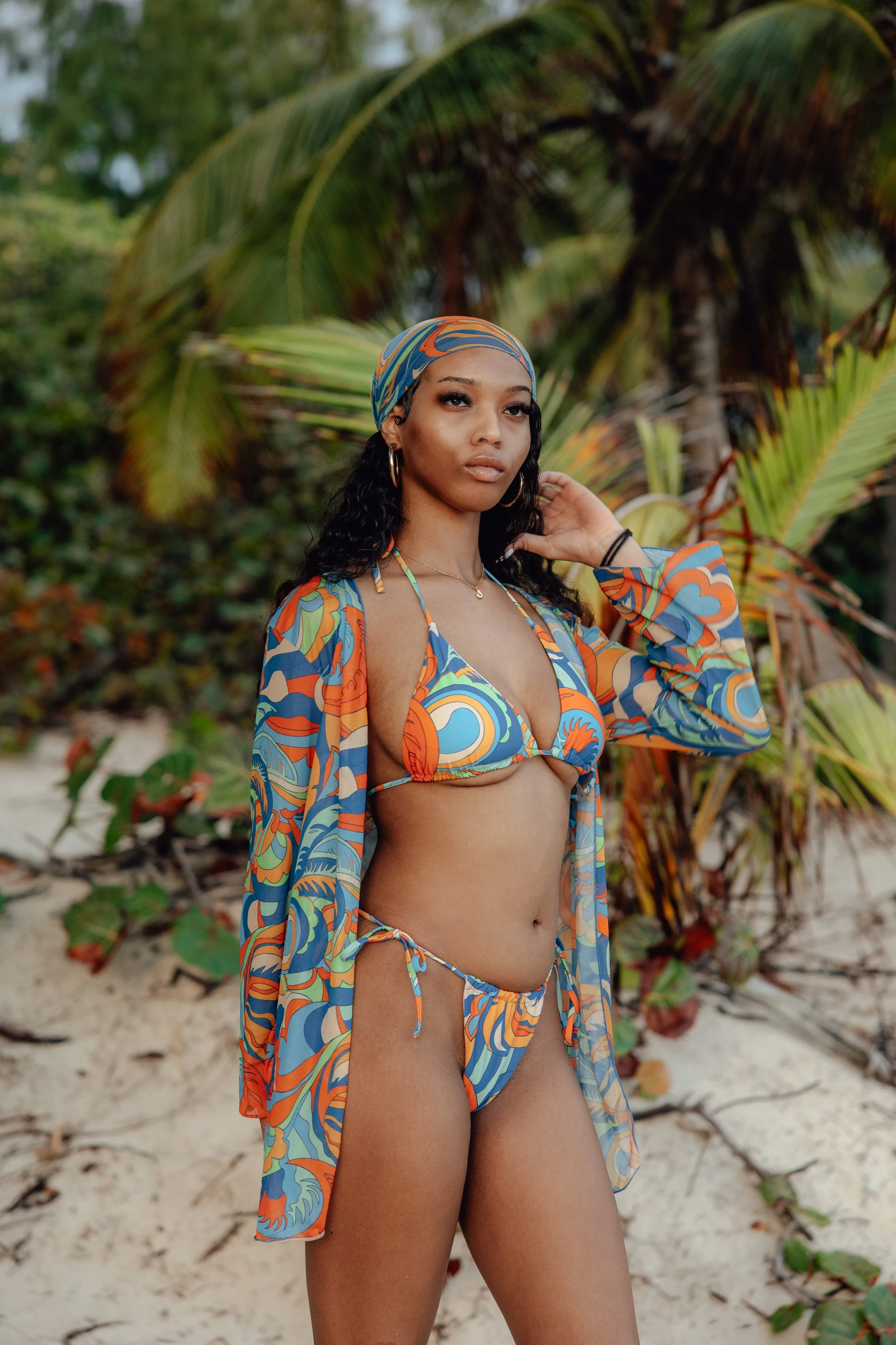 A woman with long dark curly hair wearing a colorful bikini and matching cover-up, standing on a sandy beach with tropical plants and trees in the background.