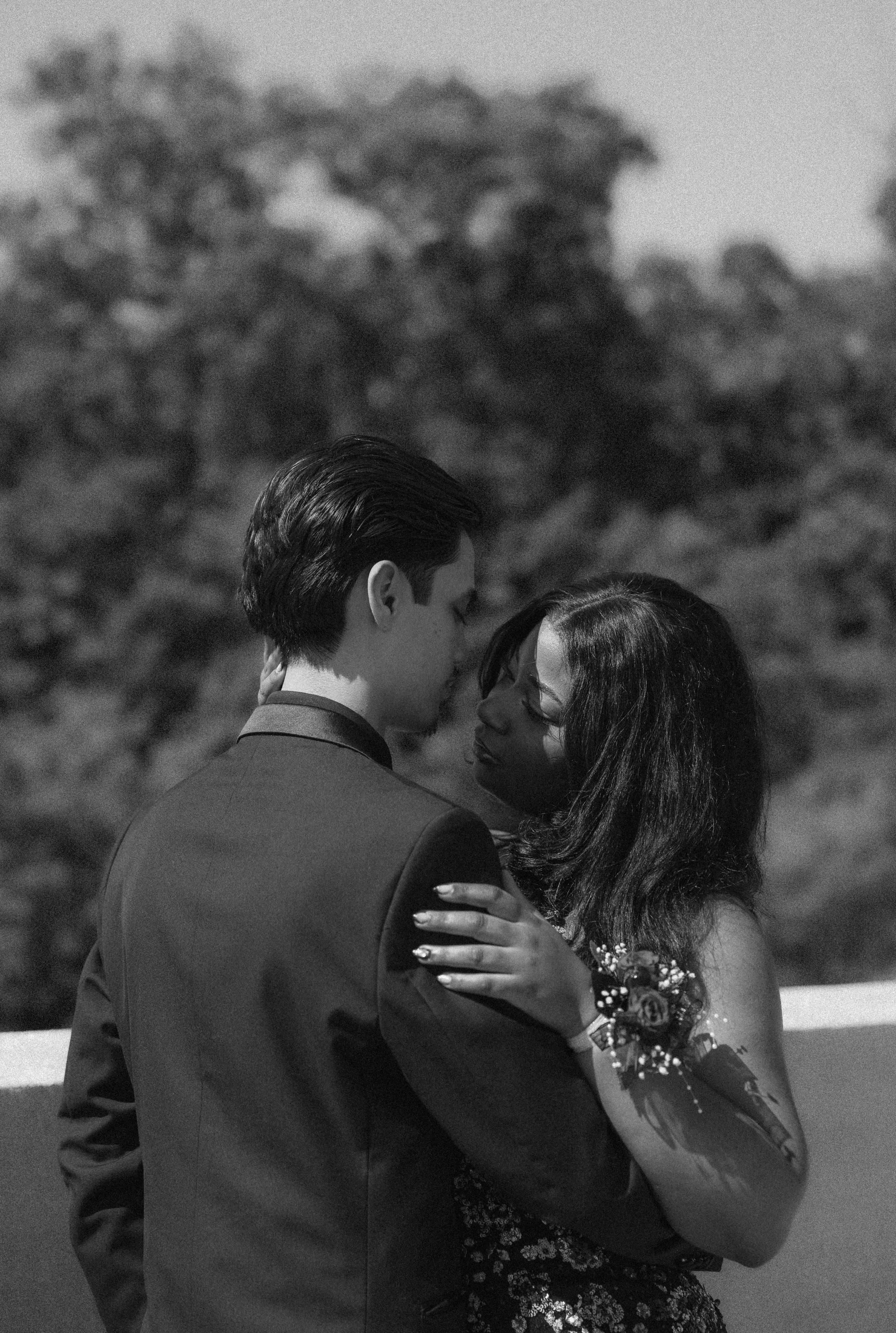 A black and white photo of a couple dancing closely outdoors, with the man wearing a suit and the woman wearing a dress with floral details. The woman also has flowers in her hair and a tattoo on her arm.