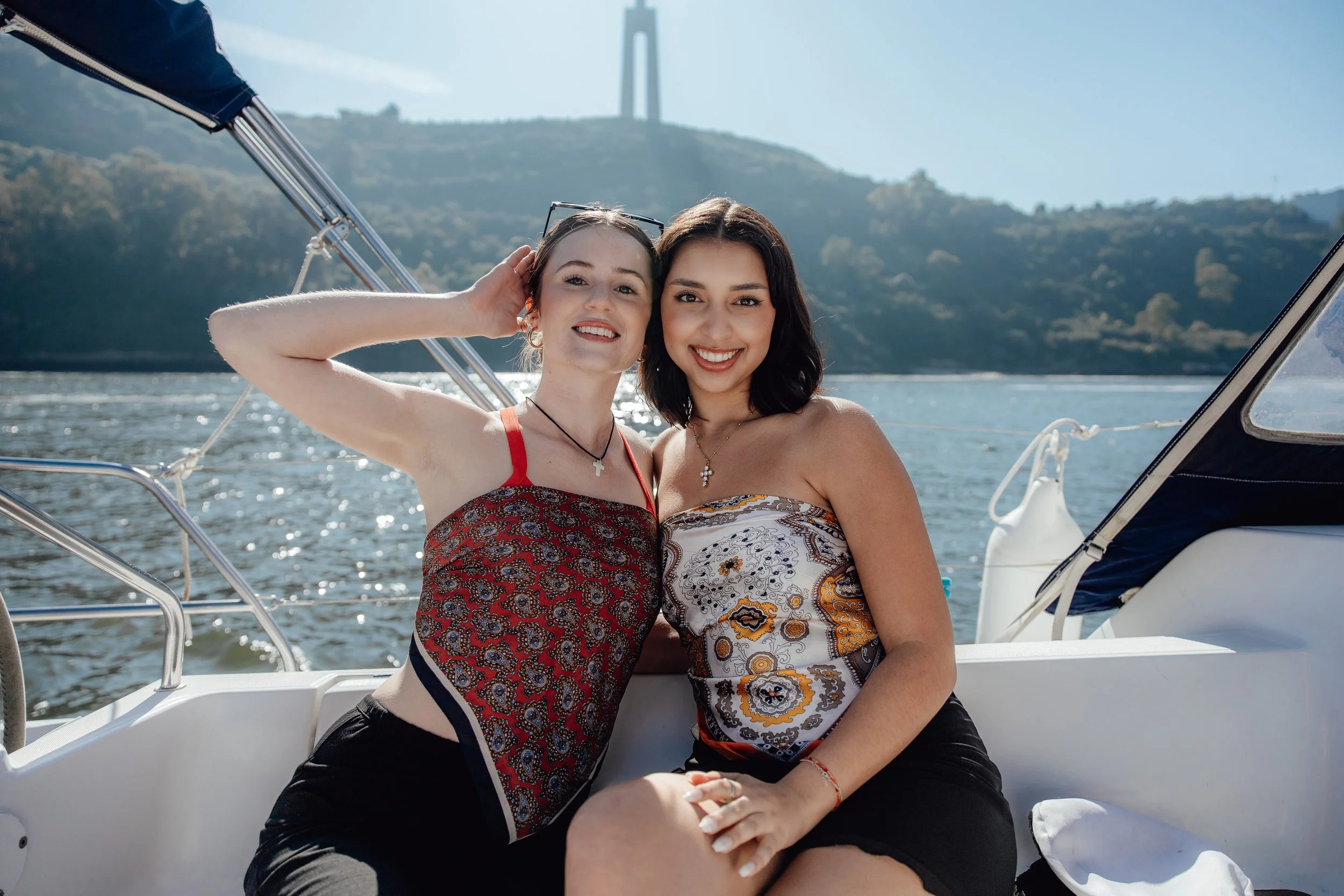 Two young women sitting on a boat, smiling at the camera, with water and hills in the background and a tall structure on the hill.