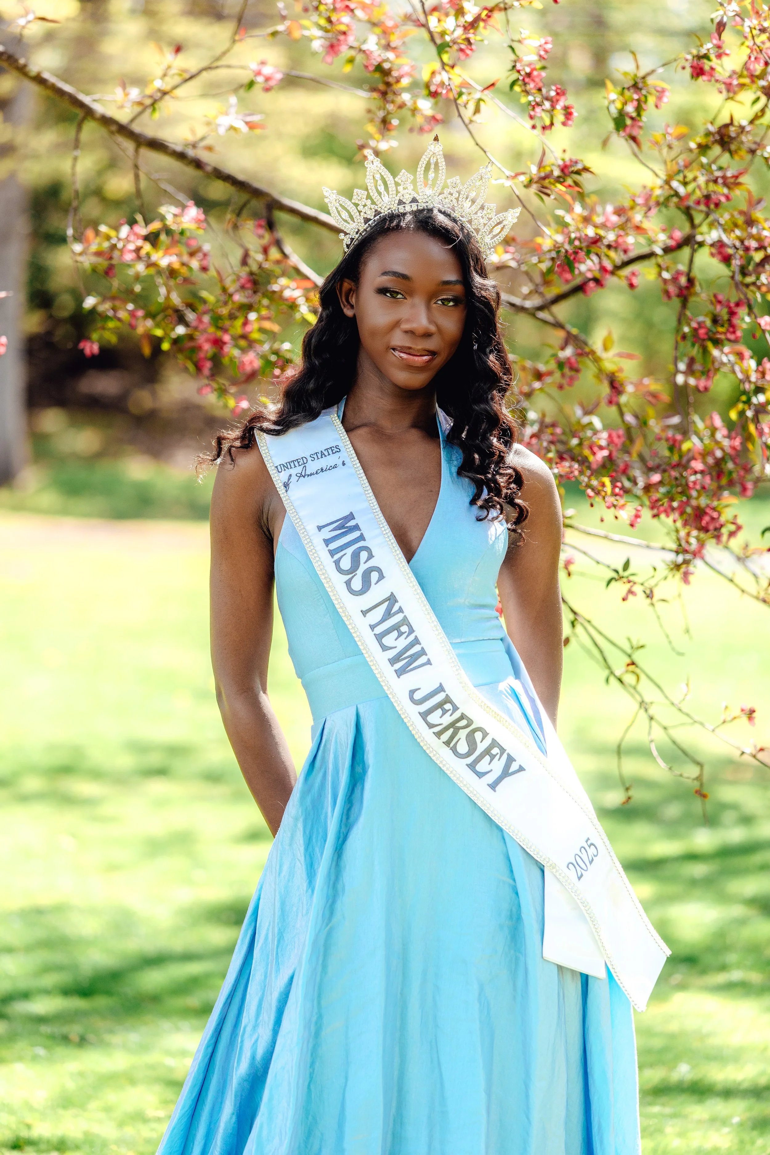 A woman in a blue dress wearing a silver crown with gemstone accents, standing outdoors in front of blooming pink flowers and green foliage, with a sash that reads "Miss New Jersey 2025."