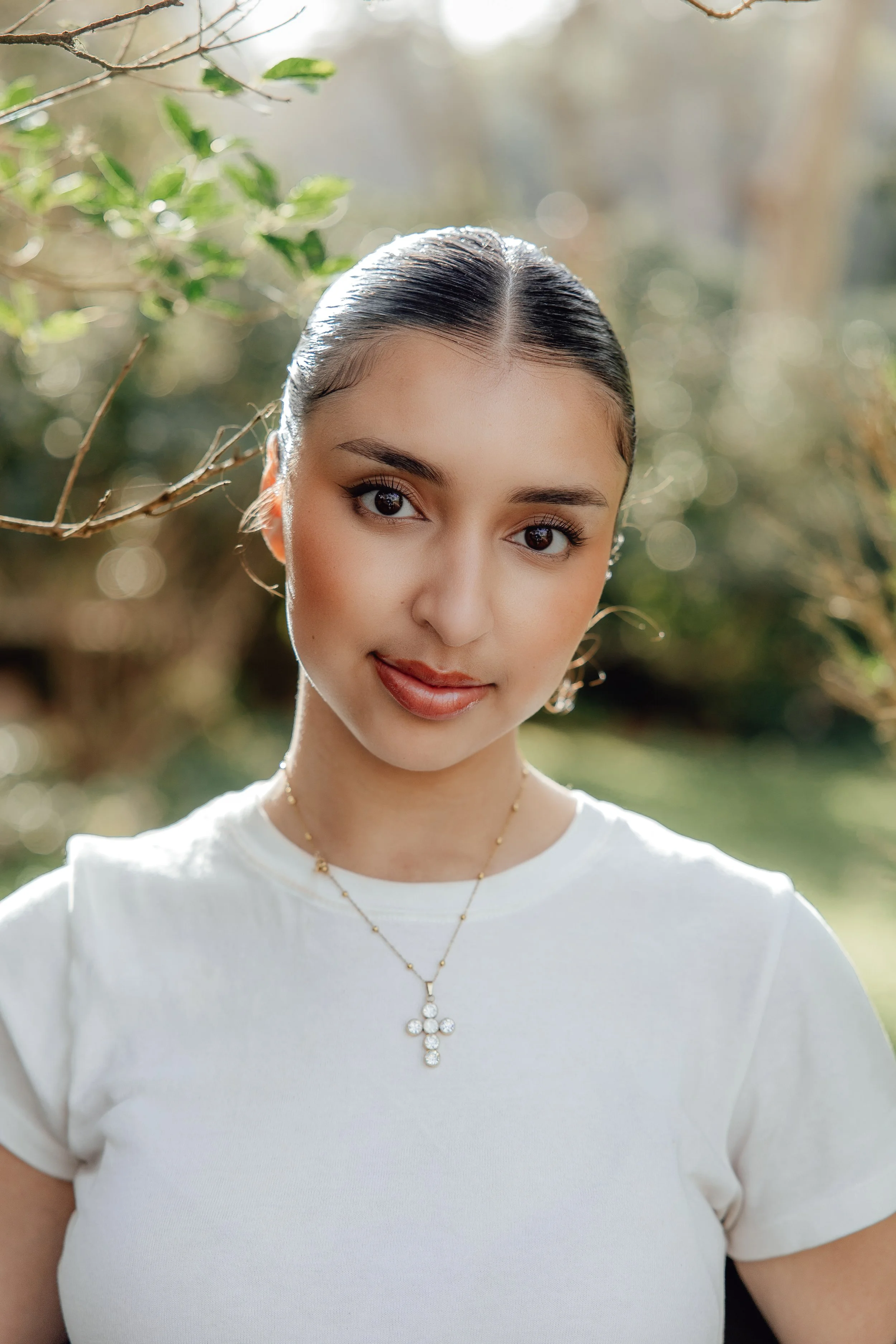Close-up of a young woman with dark hair slicked back, wearing a white top and gold jewelry, outdoors in natural light with a blurred background of green foliage.