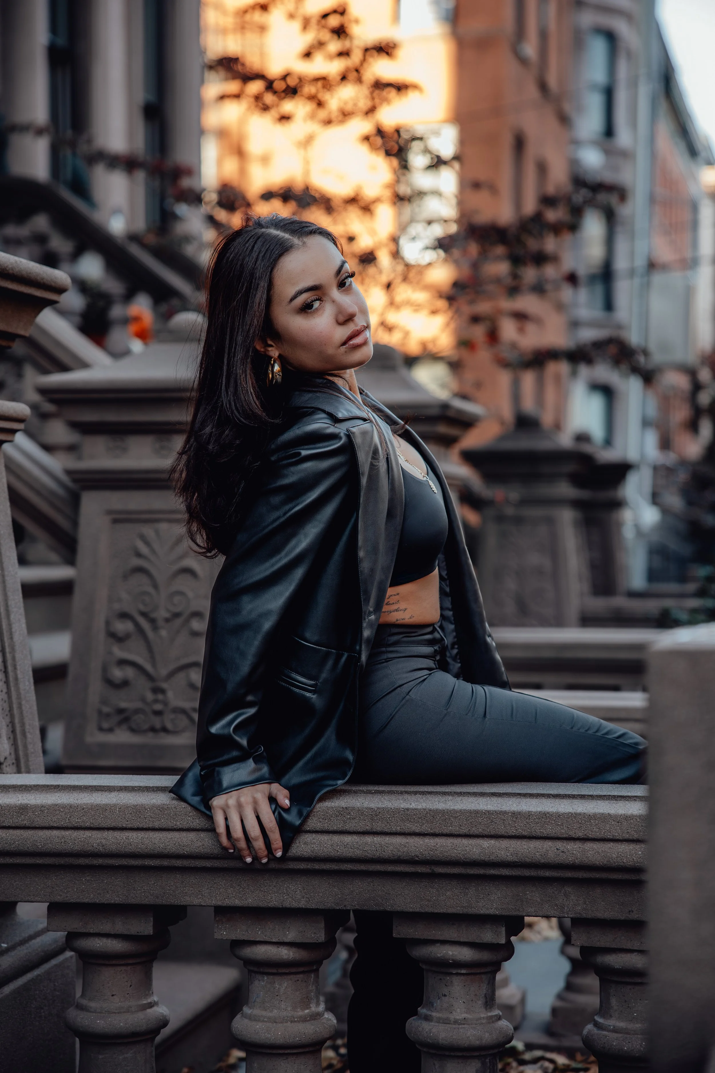 A woman with dark hair and hoop earrings dressed in black leather pants and a leather jacket posing on a stone balustrade in front of an urban brownstone building during sunset.