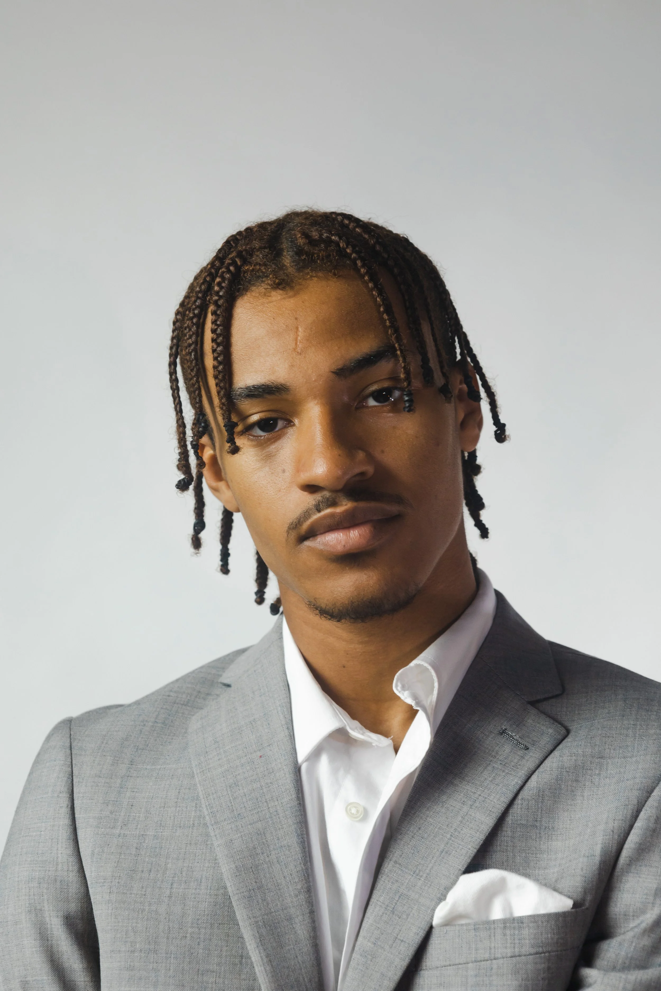 A young man with braided hair wearing a gray suit with a white shirt and a pocket square, against a plain gray background.