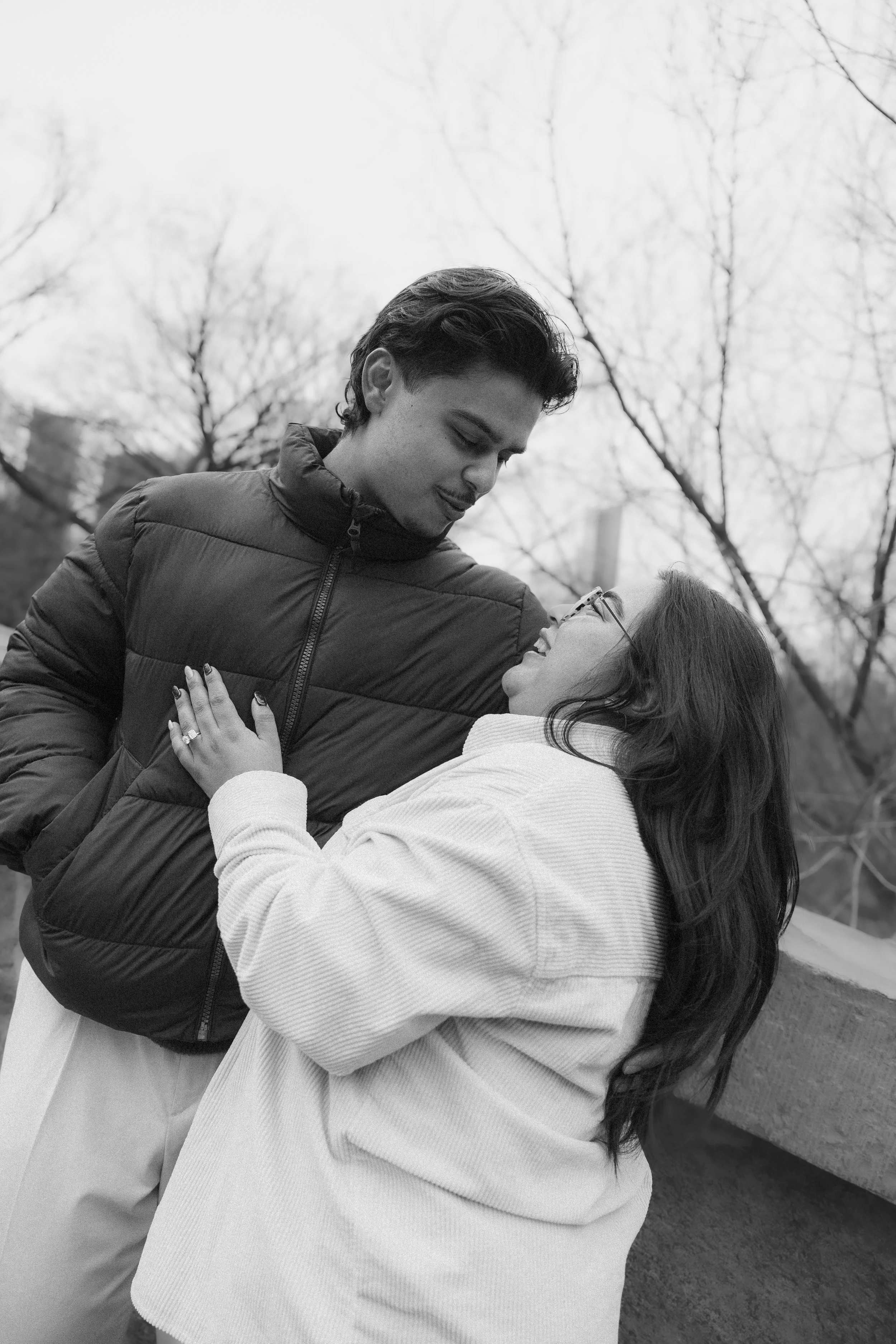 A young man and woman are outdoors, embracing and looking at each other lovingly, with leafless trees in the background.