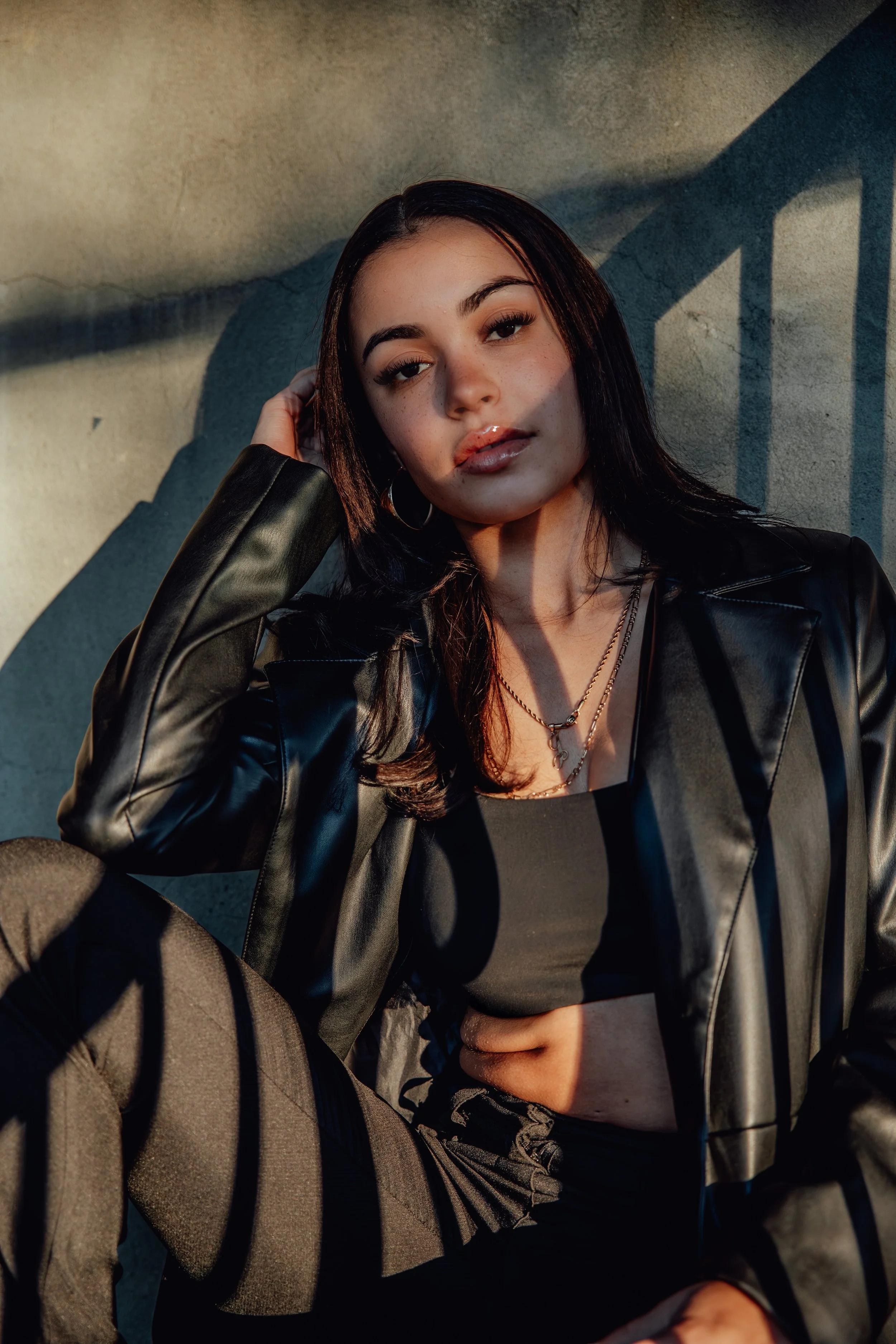A young woman with dark hair, wearing a black leather jacket and layered necklaces, sitting outdoors against a wall with sunlight casting shadows, gazing at the camera.