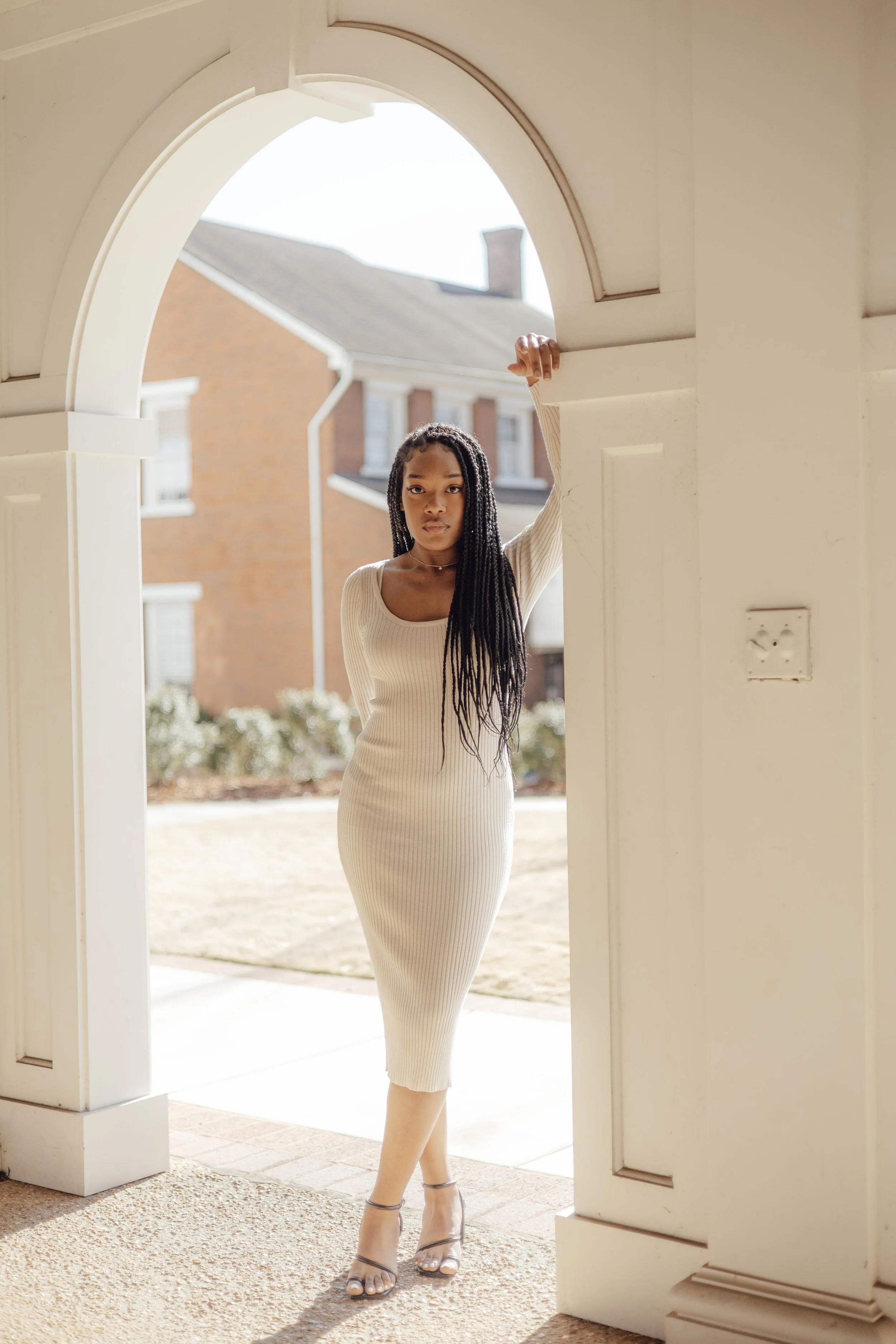 A woman in a fitted beige ribbed dress standing under an archway, posing with one hand resting on the arch, with a background of a residential neighborhood and clear sky. She has long braided hair and is wearing high-heeled sandals.