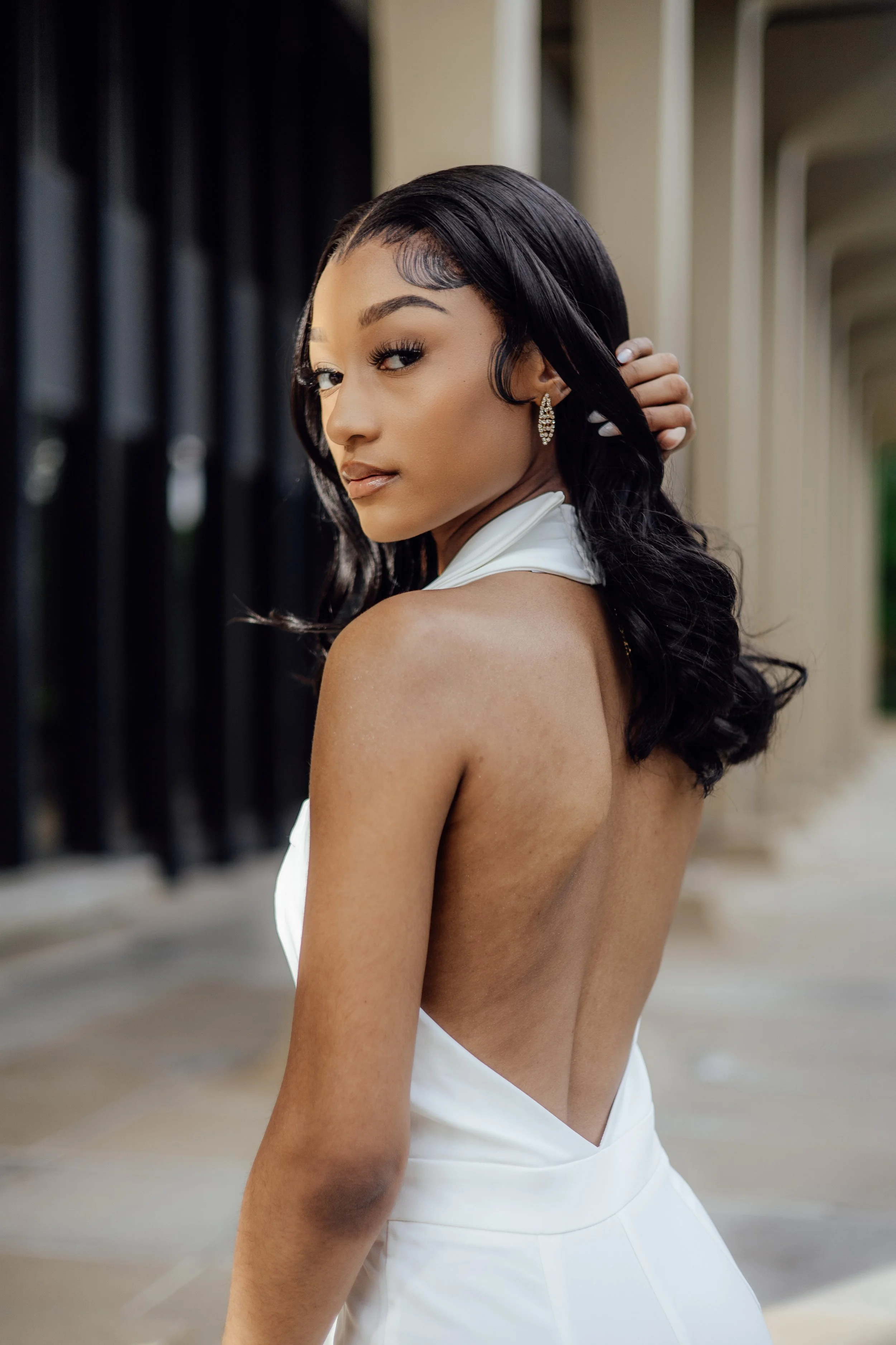 A woman with long black hair styled in loose waves, wearing a white backless dress, and silver earrings, standing outdoors near an architectural structure with columns.