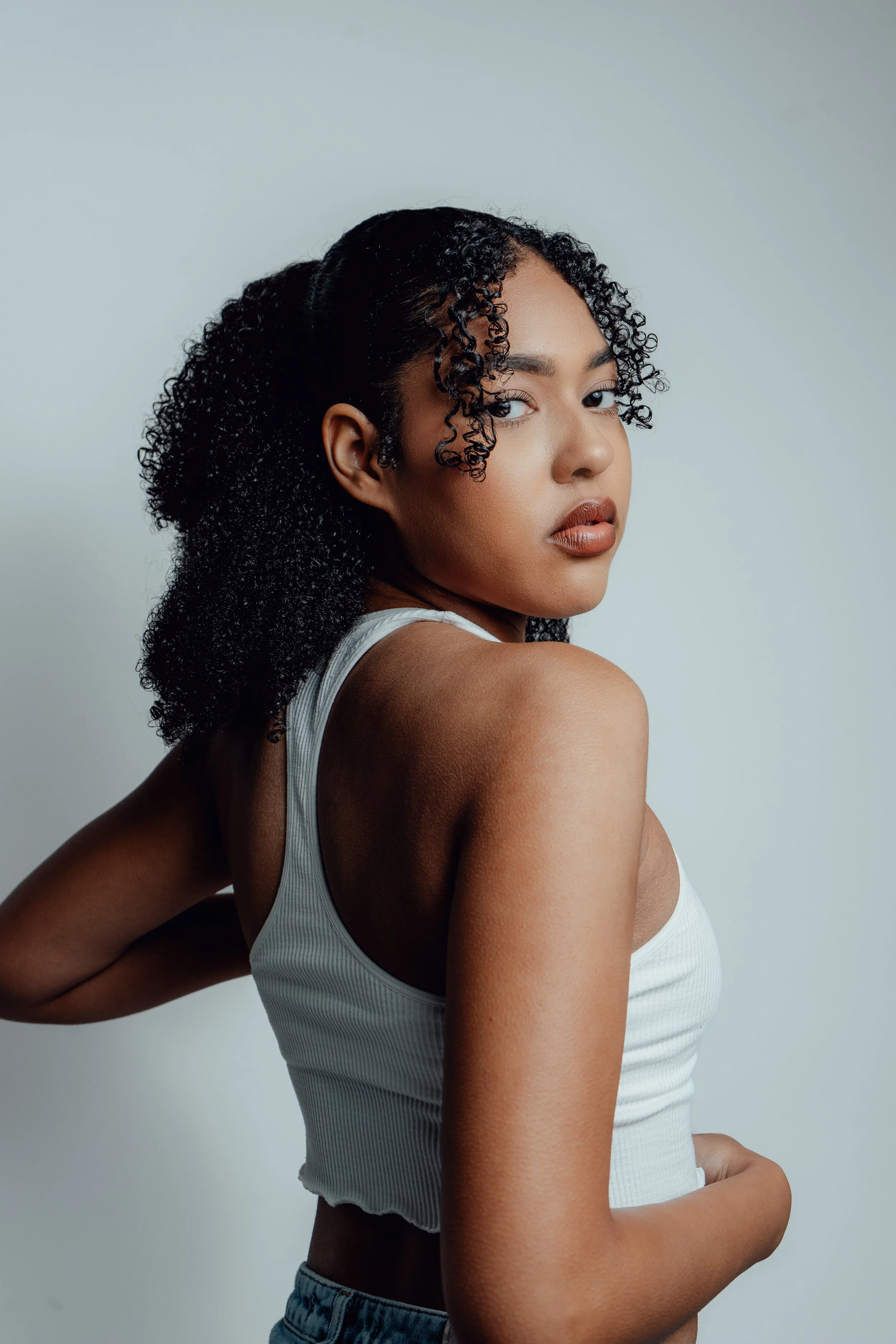A young woman with curly black hair looking over her shoulder at the camera, wearing a white tank top and jeans, with a neutral background.