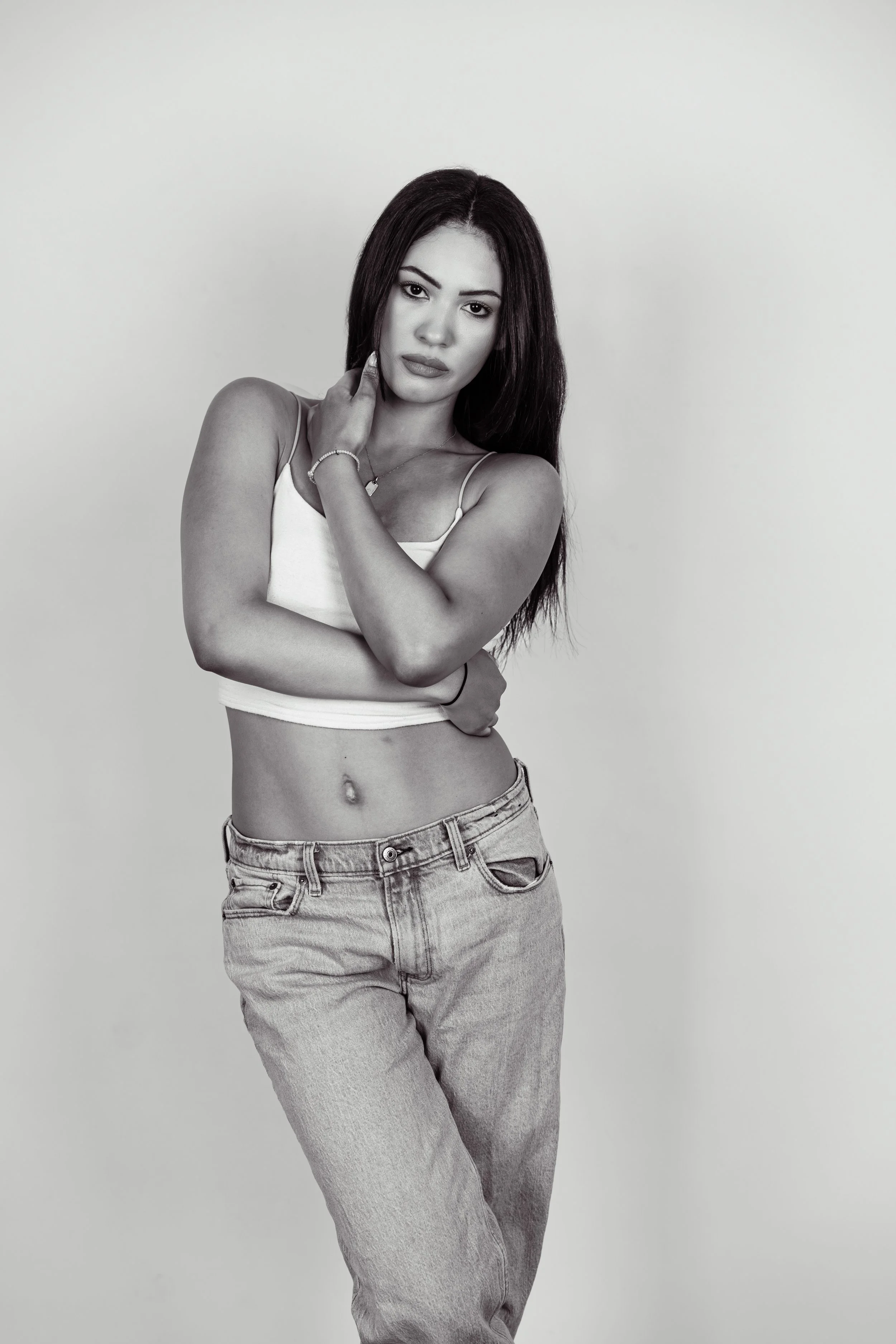 Black and white photo of a young woman with long dark hair, wearing a spaghetti strap crop top and jeans, standing against a plain background, looking directly at the camera with one hand touching her neck.