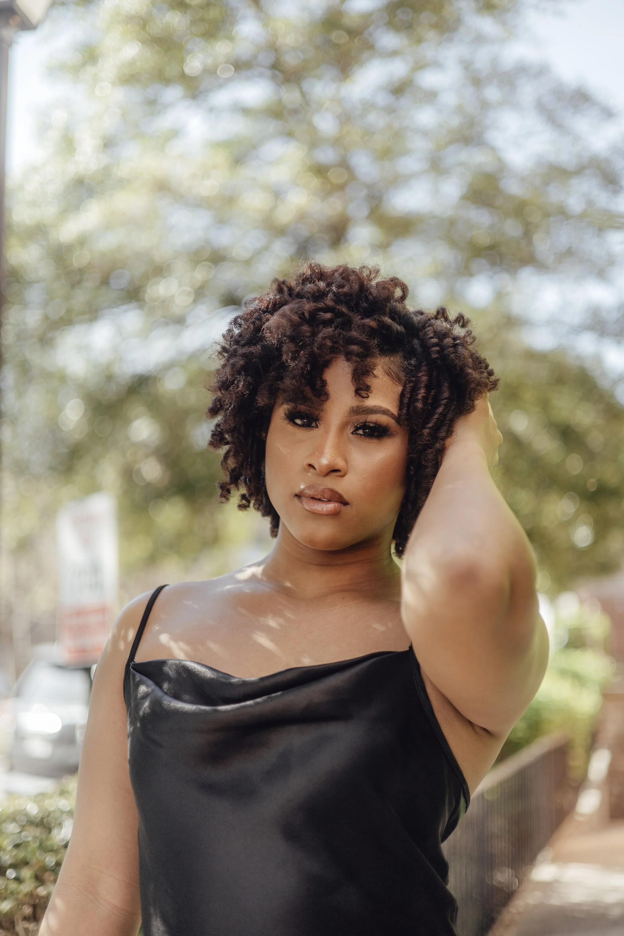 A woman with curly short hair and wearing a black satin dress is outdoors with a blurred background of trees and a street.
