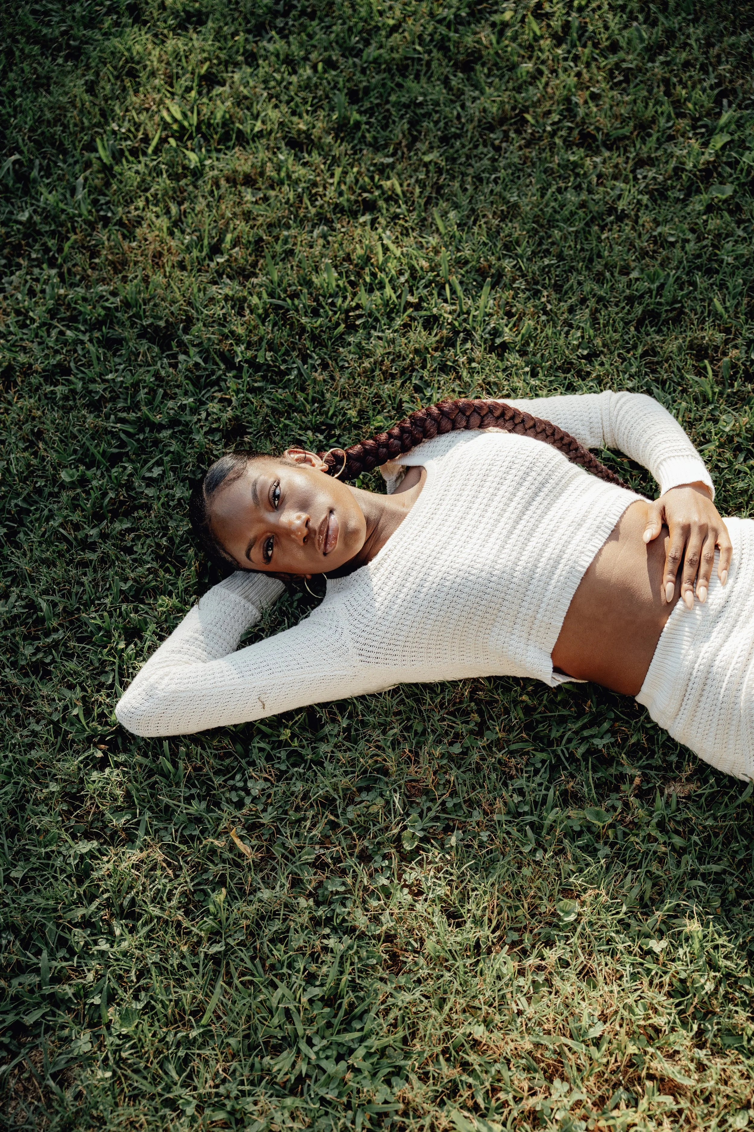 A woman with long braided hair lying on grass, wearing a white knitted sweater and gold hoop earrings, looking at the camera.