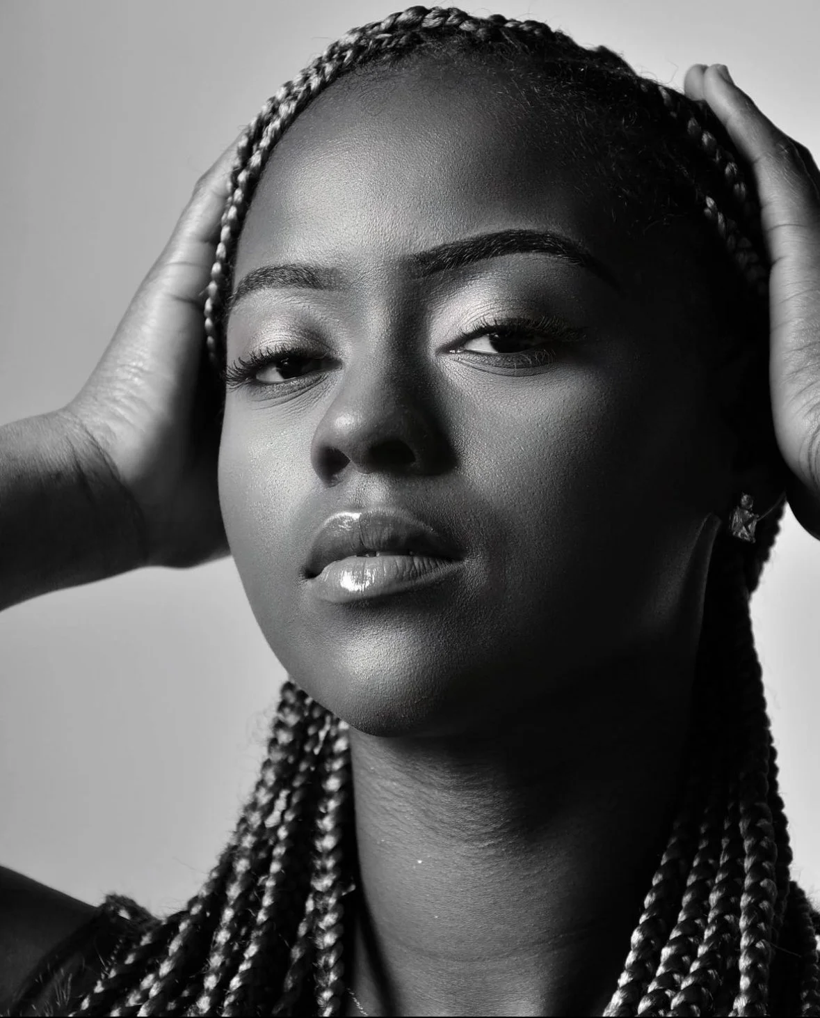 Black and white portrait of a woman with braided hair, holding her head with both hands, looking directly at the camera.