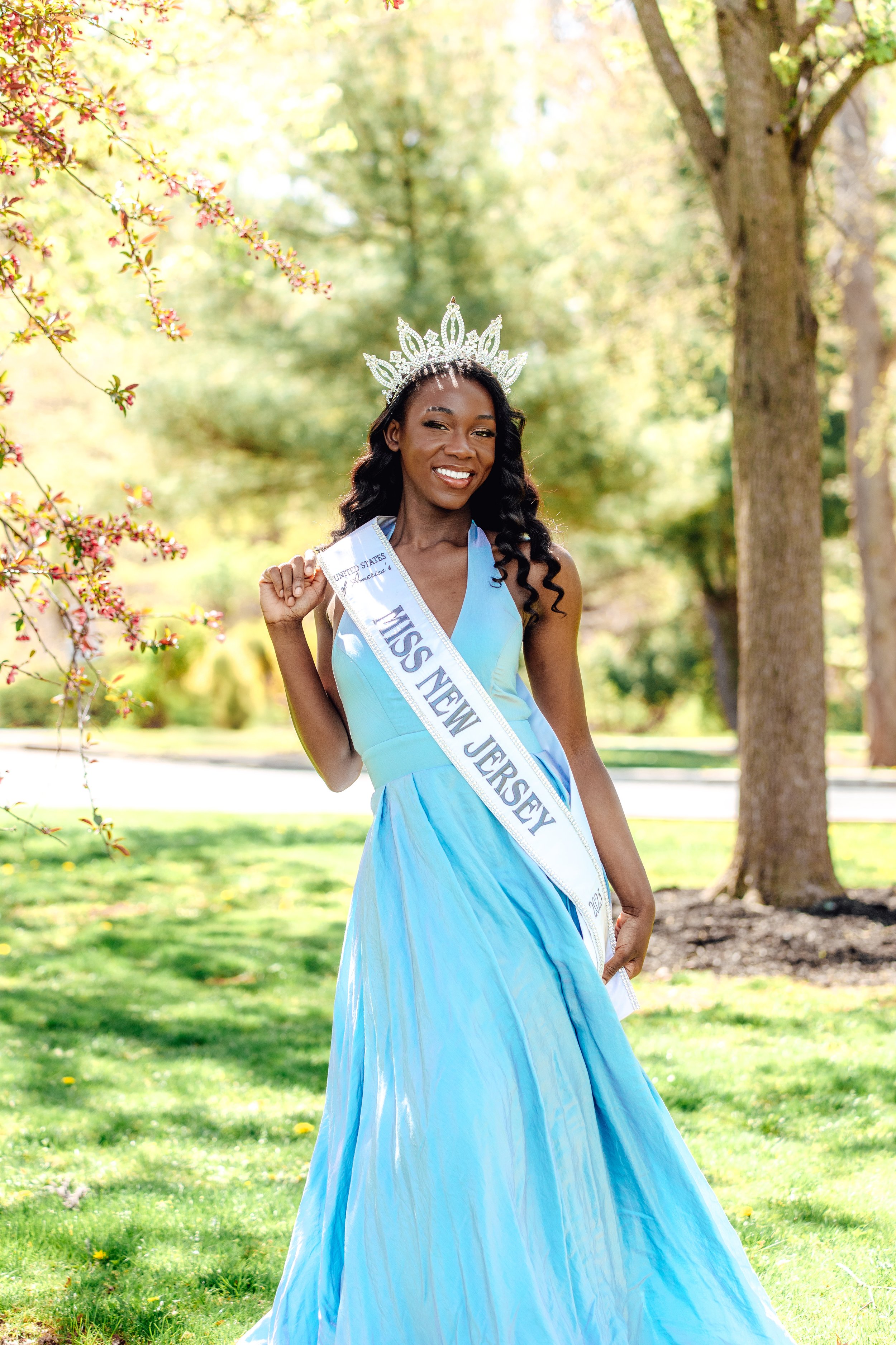 A woman wearing a light blue gown and a tiara, holding a sash that reads 'Miss New Jersey,' standing outdoors in a park with green grass, trees, and pink flowering branches in the background.