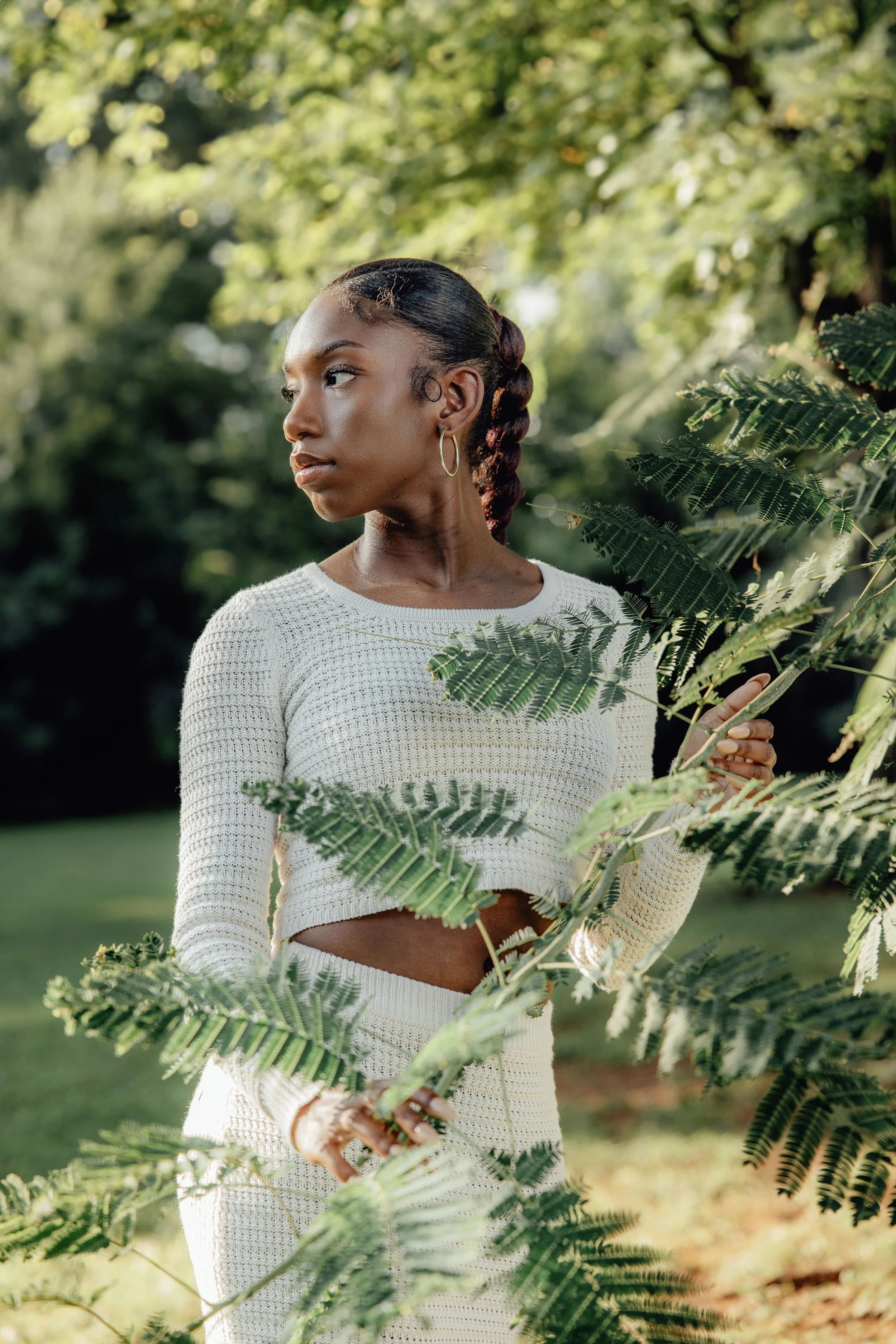A young woman with dark skin and braided hair tied in a low ponytail standing outdoors among green foliage, wearing a white textured long-sleeve top and hoop earrings, with sunlight filtering through trees in the background.