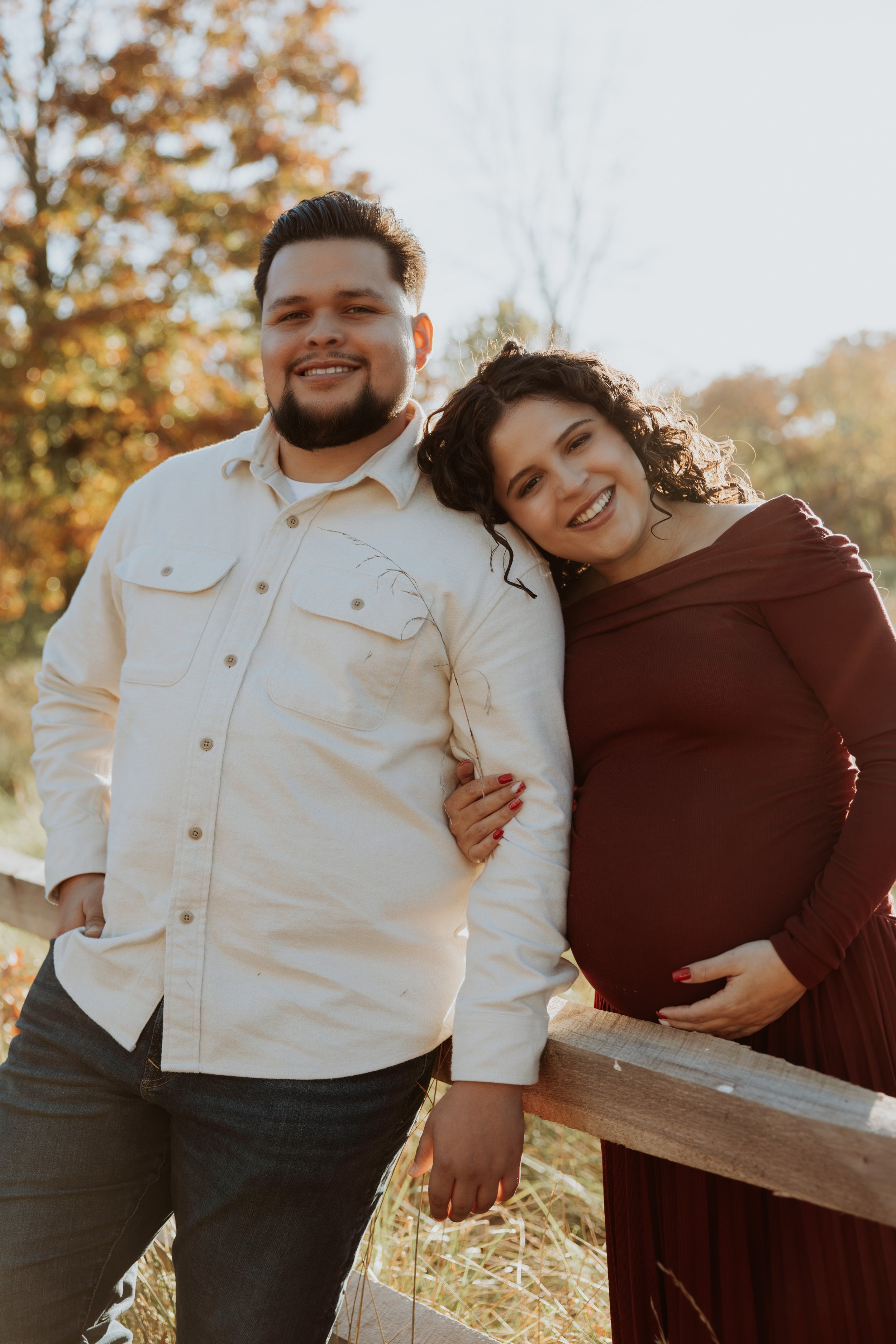 A smiling man with a beard and a woman with curly hair pose together outdoors during fall, with trees and golden leaves in the background. The woman is holding her pregnant belly.