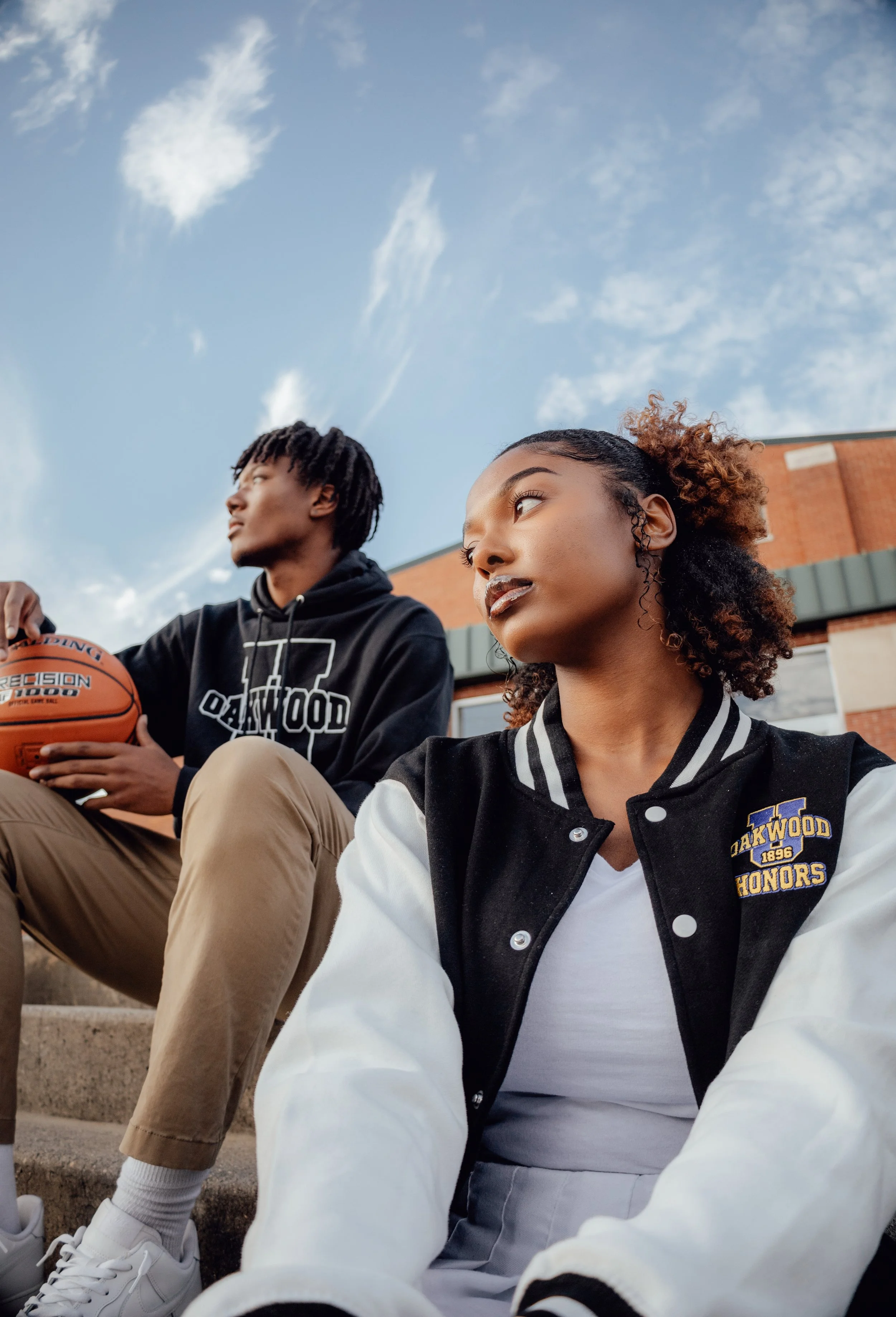 Two teenagers sitting on outdoor steps, a boy holding a basketball, and a girl wearing a jacket with a school logo, against a blue sky with clouds.