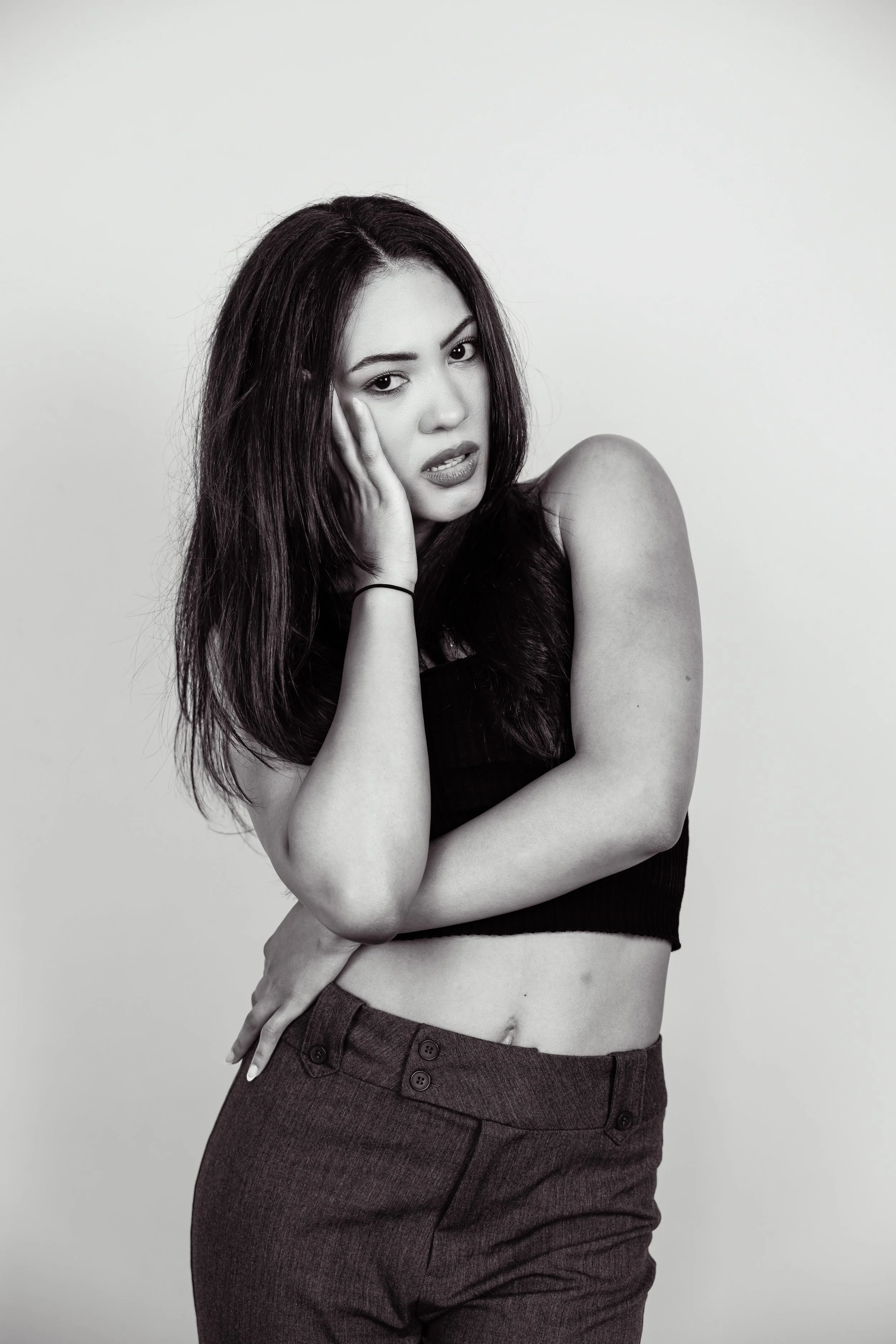 Black and white photo of a young woman with long dark hair, wearing a sleeveless top and high-waisted pants, posing with her hand on her face and a contemplative expression.