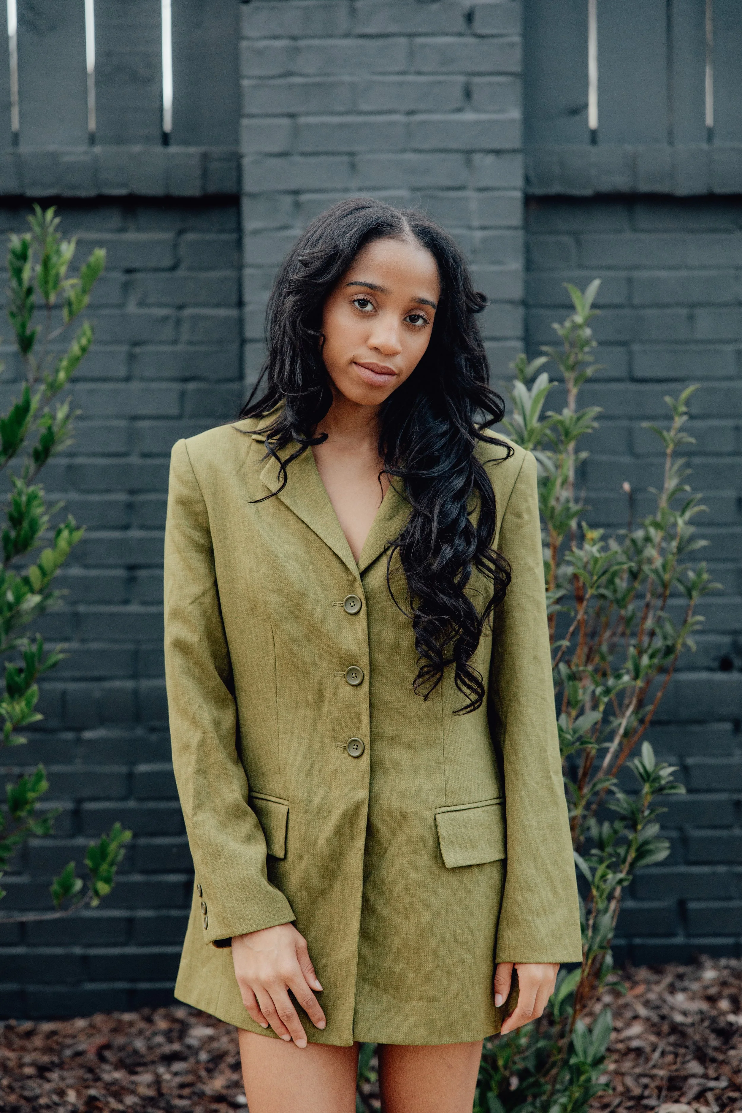 A woman with long black hair and light brown skin standing outdoors in front of a black brick wall and some green shrubs, wearing an olive green blazer and matching shorts.