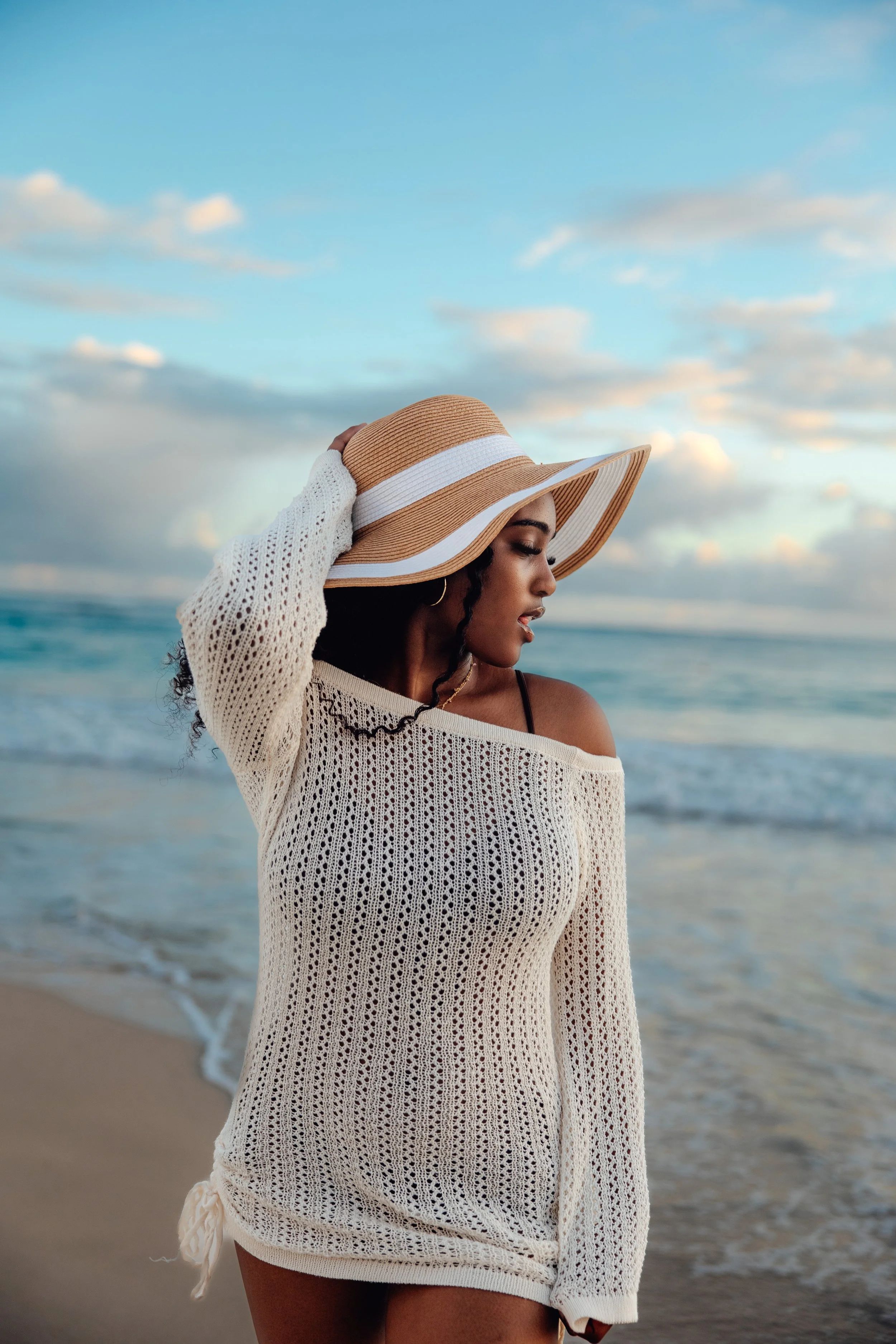 A woman with dark curly hair wearing a large straw sunhat and a white knitted off-shoulder dress standing on the beach with the ocean and cloudy sky in the background.