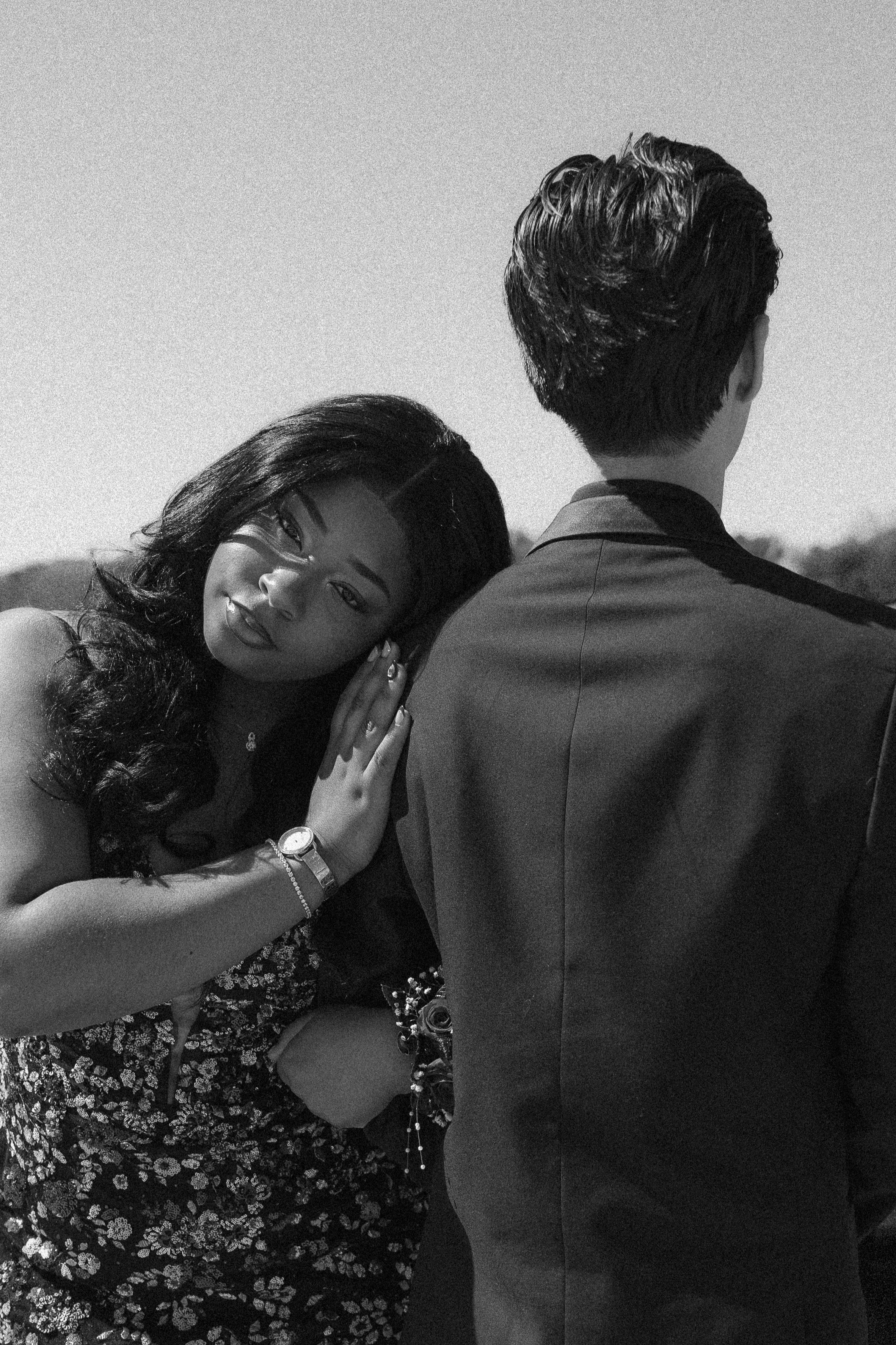 A woman with long dark hair and a patterned dress leaning her head on the shoulder of a man with short dark hair, wearing a suit, against a clear sky.
