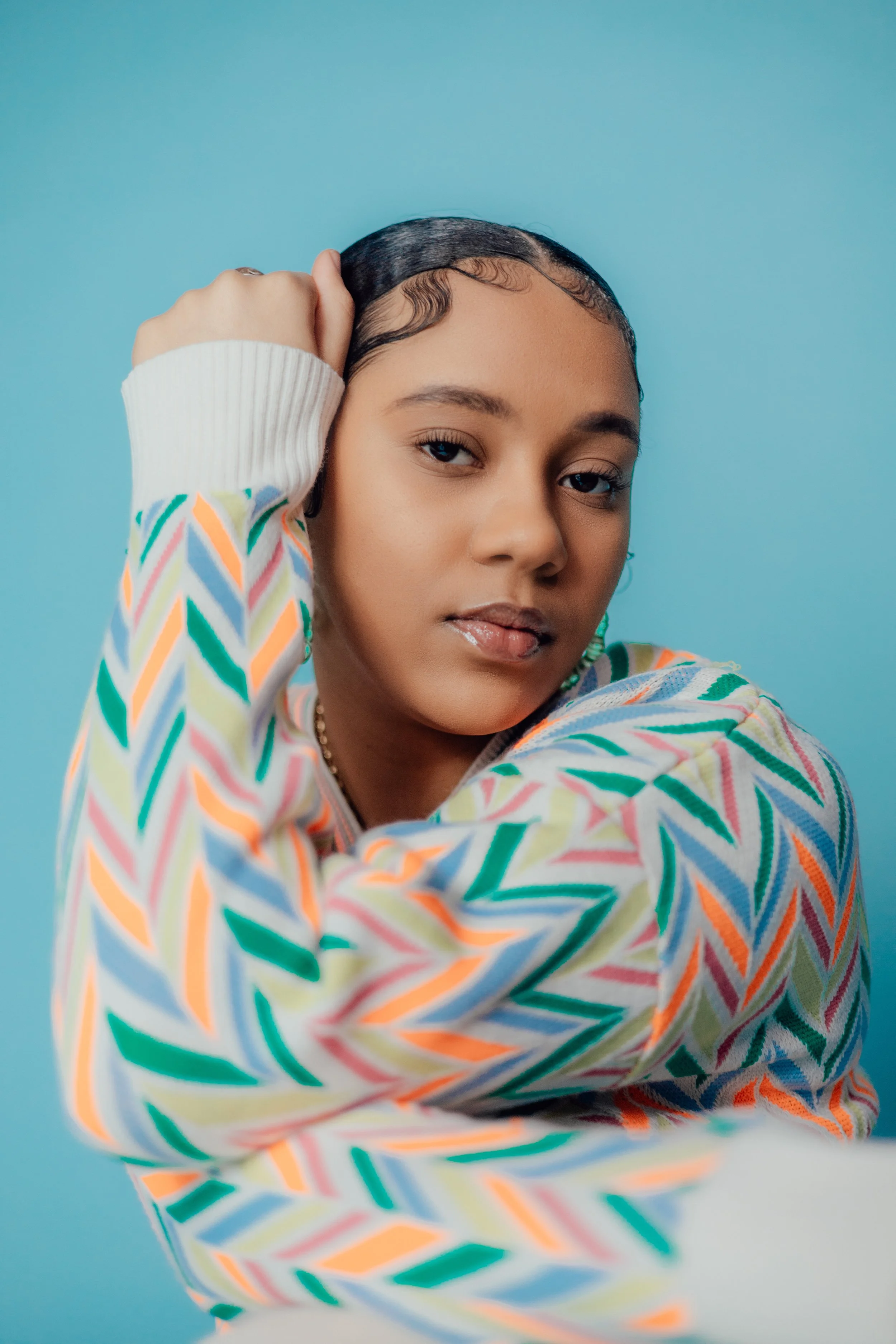 Close-up portrait of a young woman with curly hair, wearing a colorful striped sweater, against a light blue background, with her hand resting on her head, looking at the camera.