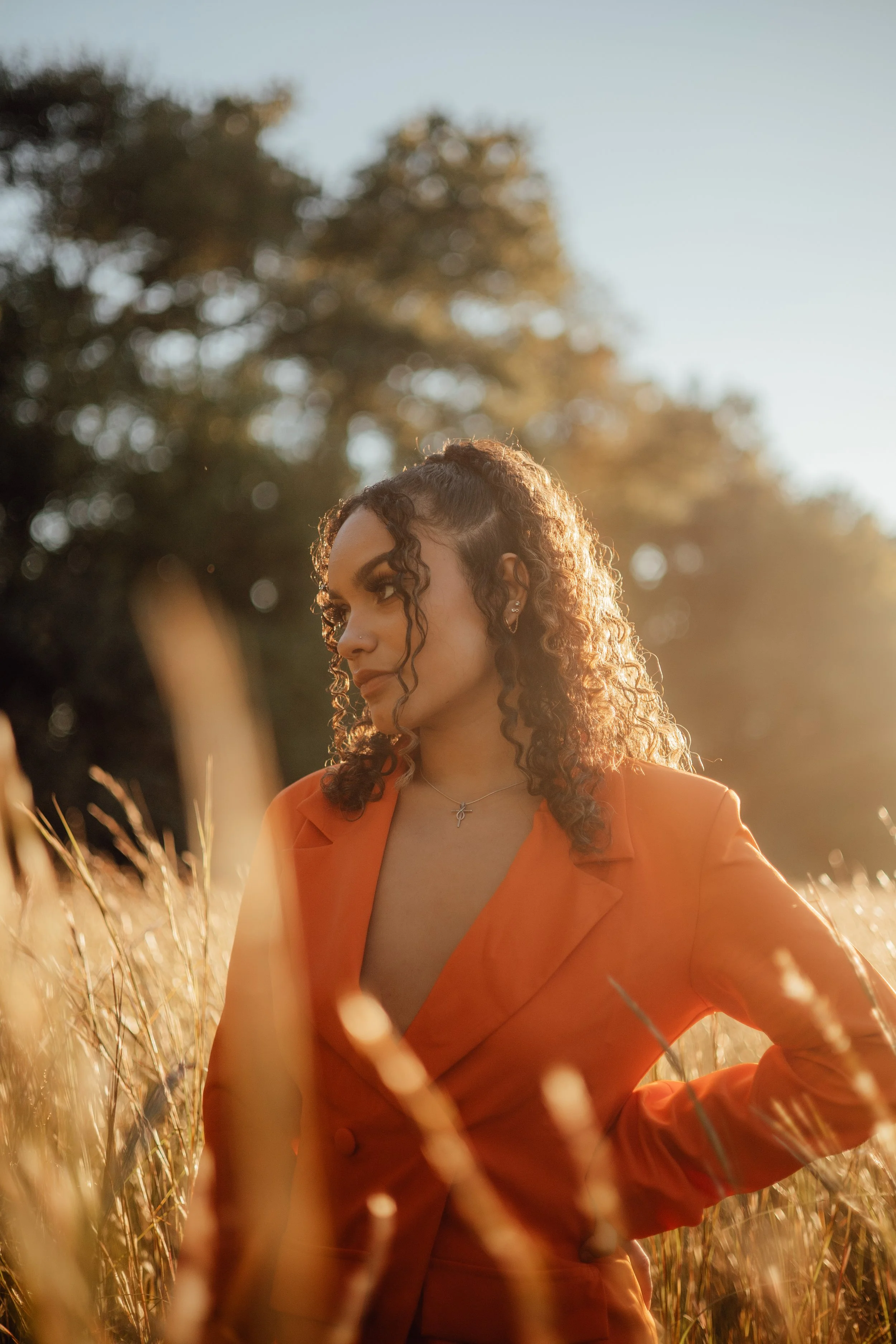 A woman with curly hair wearing an orange blazer sits in a field of tall grasses during sunset.