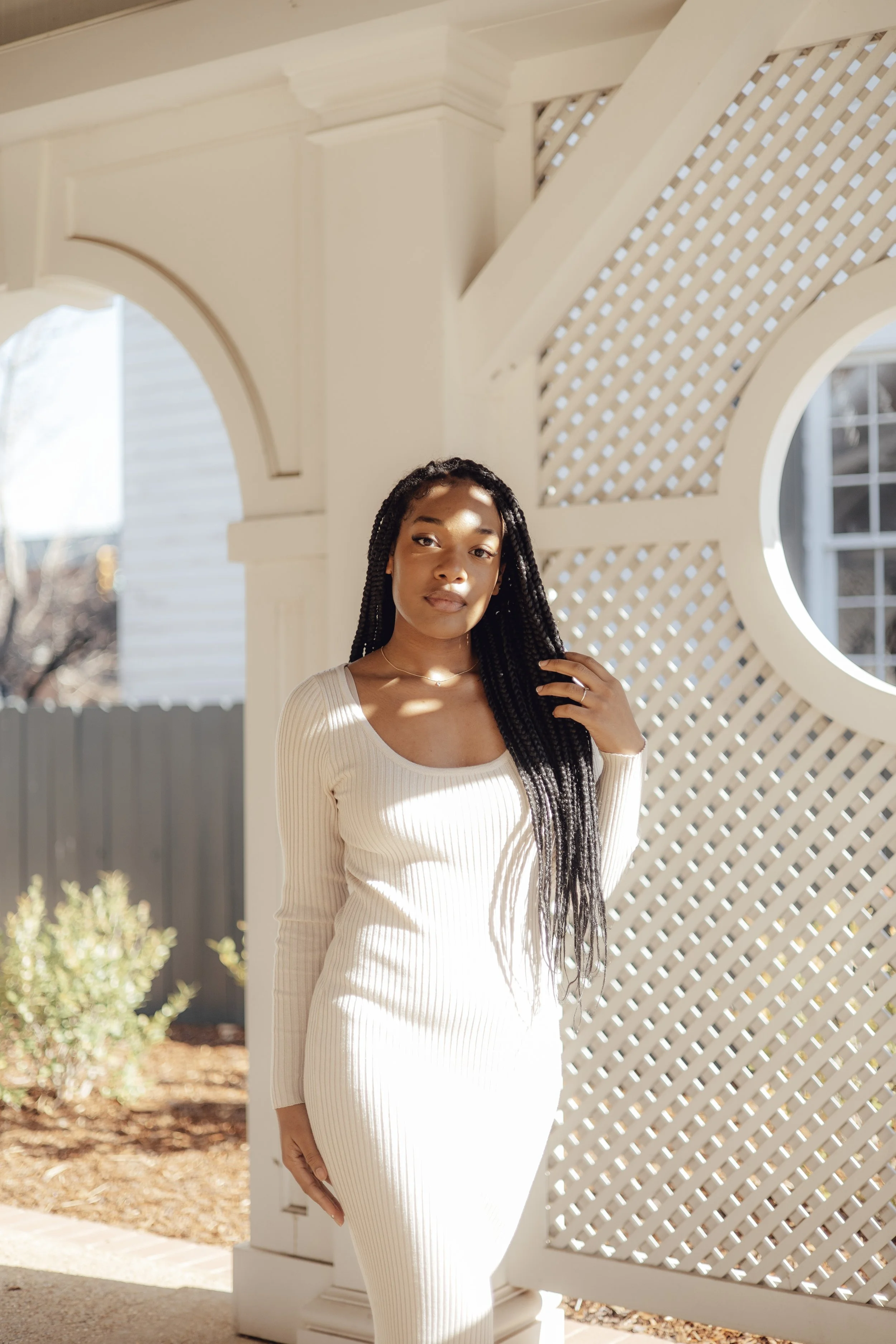 A woman with long braided hair stands outdoors near a white decorative lattice and archway, wearing a light cream ribbed knit dress, with sunlight creating shadows on her face and dress.