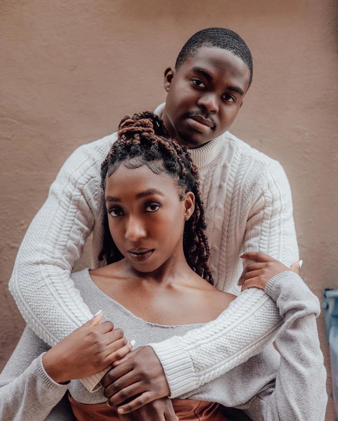 A young couple standing close together outdoors against a brown wall; the man is wearing a cream cable-knit sweater and the woman is wearing a light gray off-shoulder sweater, with her hair styled in dreadlocks.